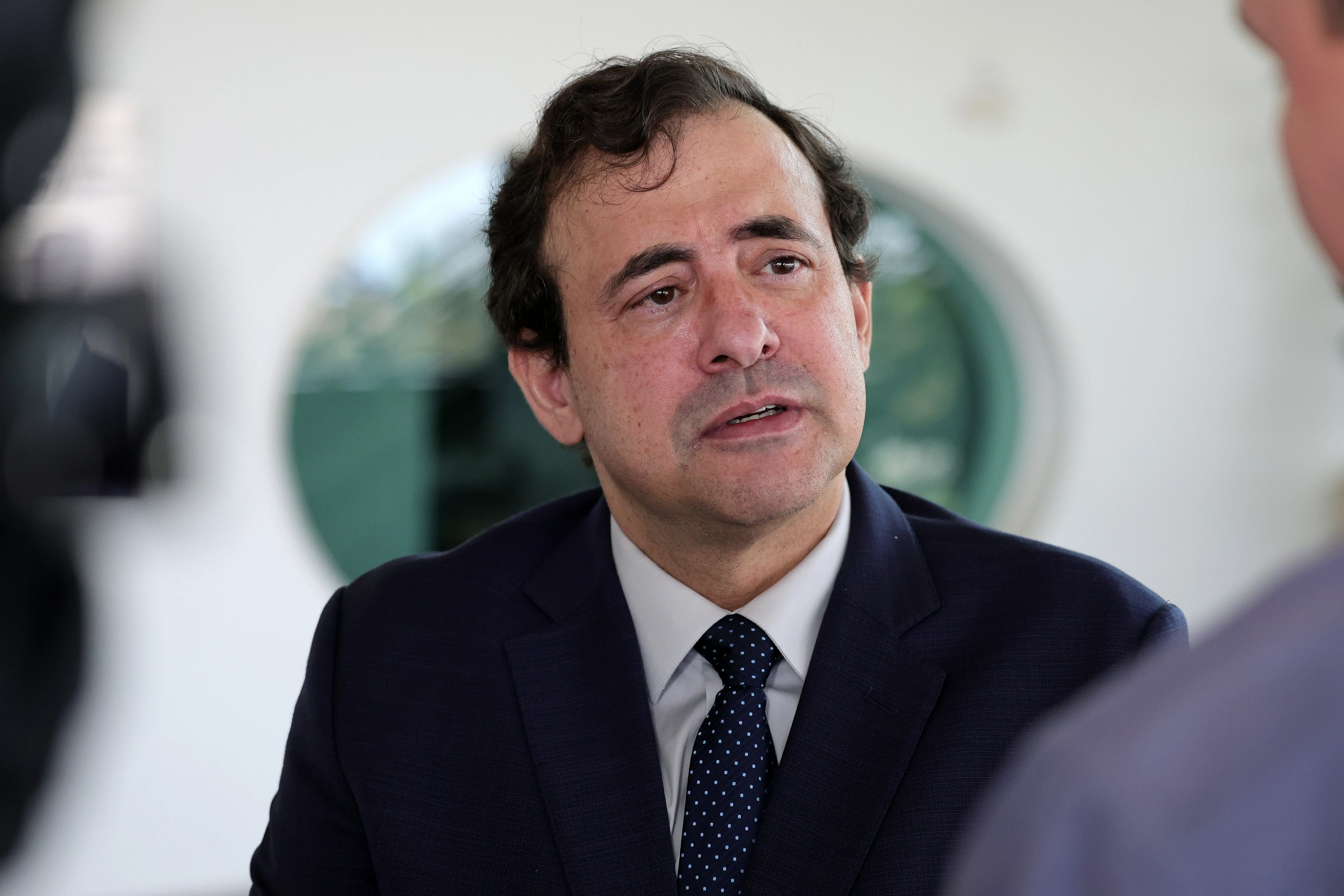 Homem de paletó azul-marinho, camisa branca e gravata escura com bolinhas fala em entrevista. Foto focada em seu rosto de perfil angular, com fundo claro e desfocado.