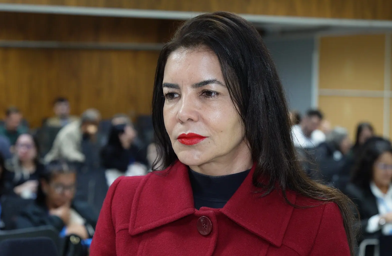 Foto horizontal que mostra a juíza Ana Paula Gomes de Freitas, do busto pra cima, olhando para o lado. Ela é uma mulher de pele parda, olhos e cabelos castanhos, usando blusa preta, batom e sobretudo vermelhos. Ao fundo, sala de aula repleta de pessoas sentadas em carteiras.