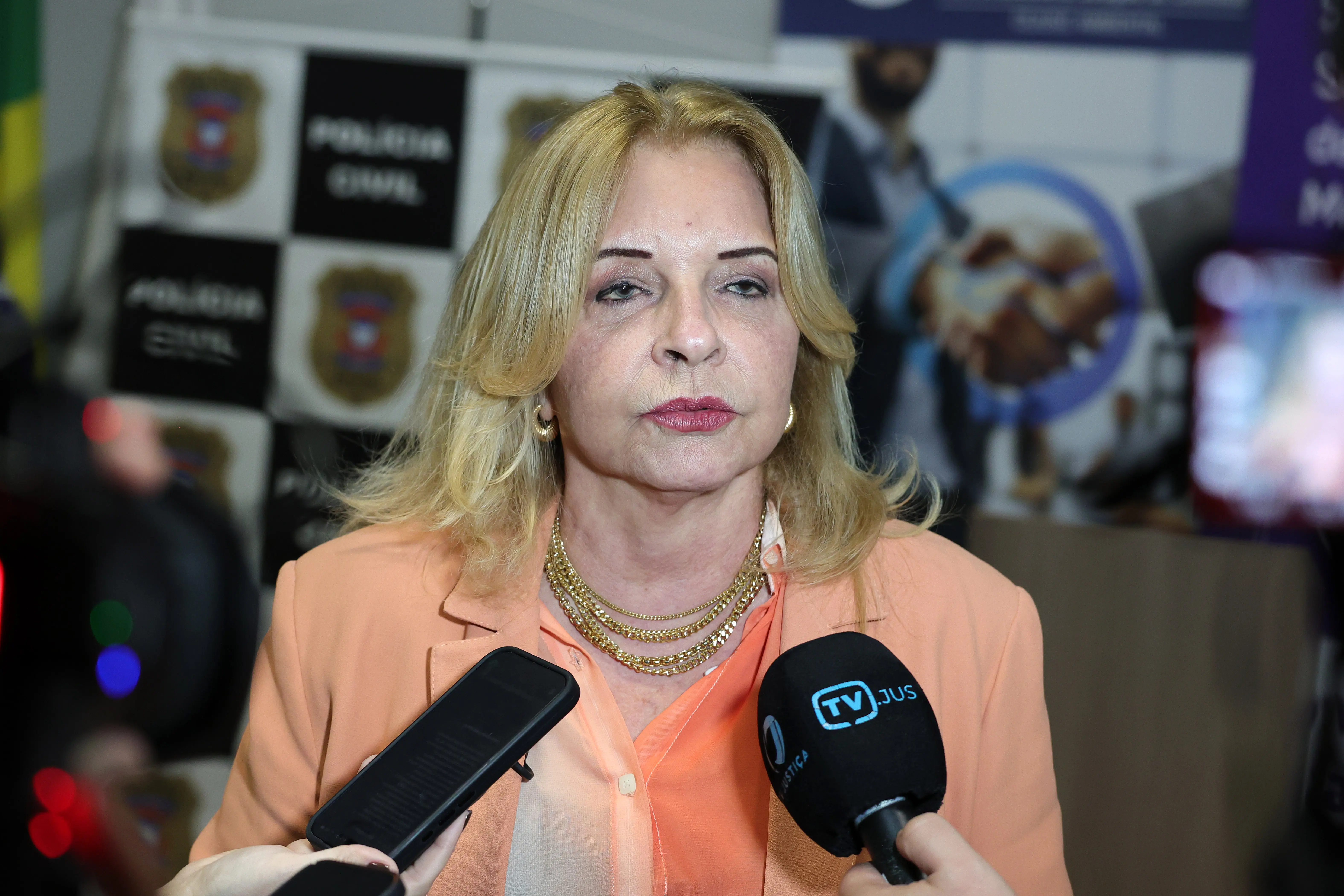 Uma mulher de cabelo loiro, blazer salmão e colares dourados concede entrevista. Em primeiro plano, duas mãos seguram microfones com a marca "TV JUS" e um smartphone direcionados a ela. Ao fundo, parede com o logotipo da Polícia Civil.