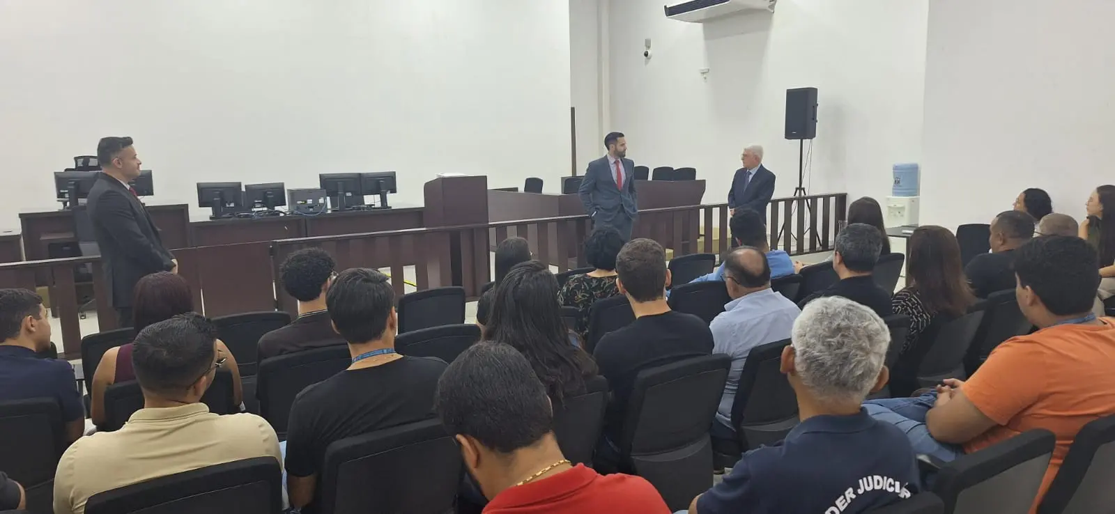 Foto do plenário do Fórum. Juízes conversam com o escrivão judicial aposentado durante a comemoração dos 44 anos da Comarca e servidores acompanham sentados.