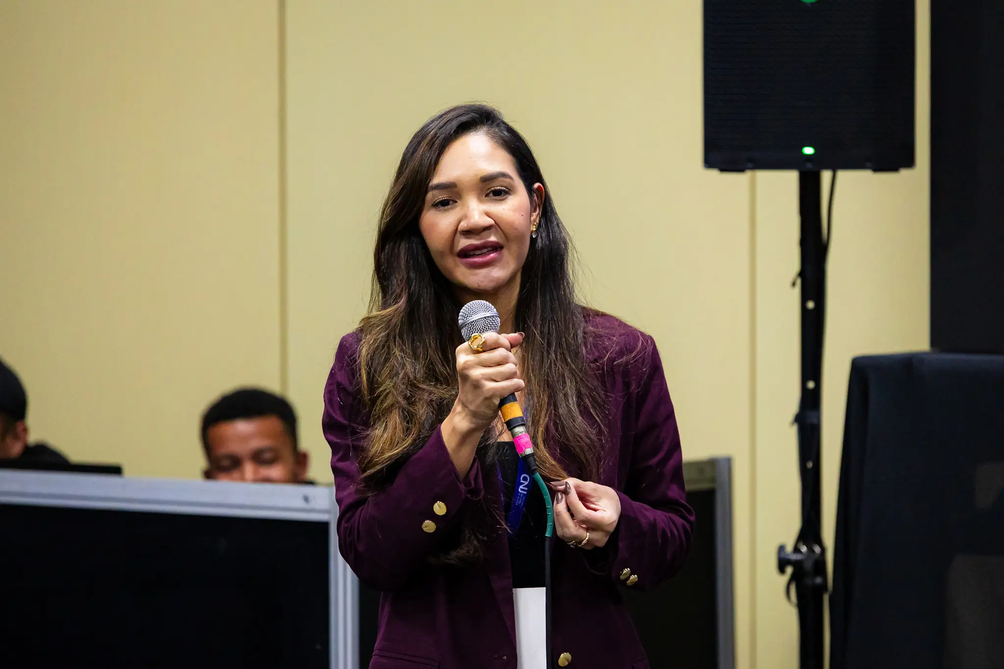 Rayandria, mulher de cabelos longos e escuros, discursa ao microfone, de blazer vinho e brincos dourados, gesticula durante apresentação em ambiente institucional.