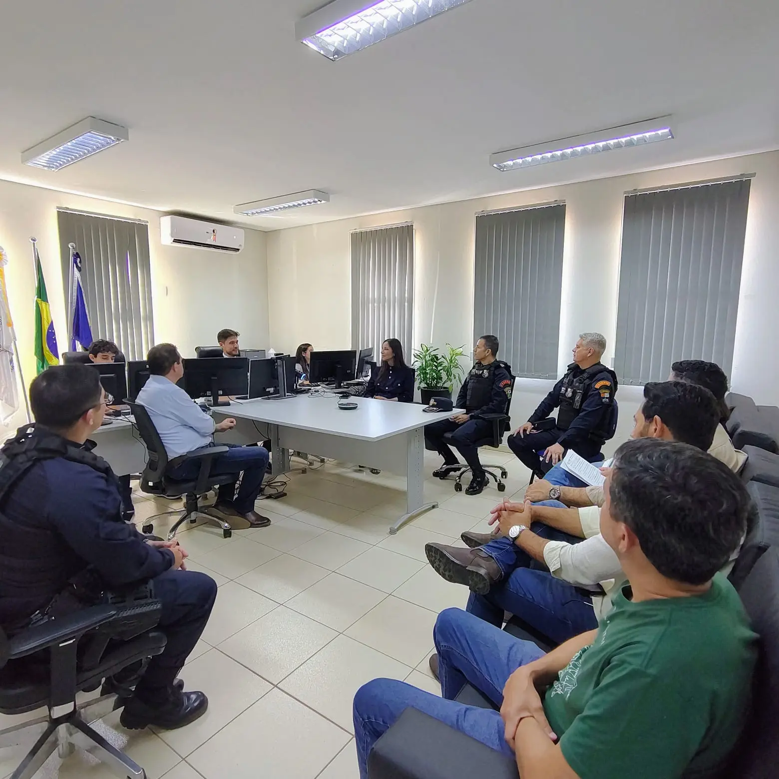 Sala de reunião com cerca de 10 pessoas ao redor de mesa com computadores. Há três policiais fardados, pessoas civis e bandeiras do Brasil ao fundo.