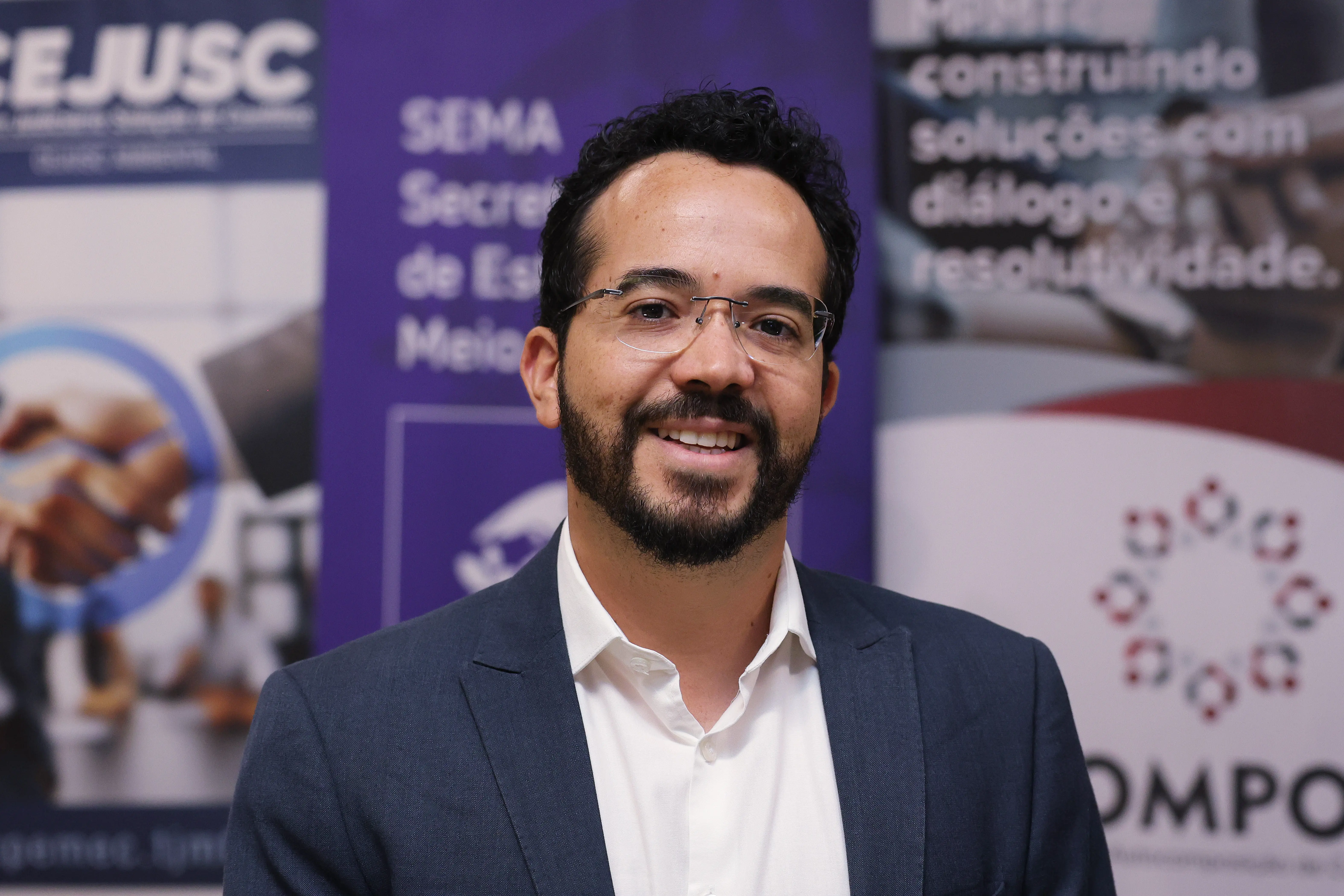 Retrato em plano médio de um homem sorrindo, de barba, óculos de grau, camisa branca e paletó azul-marinho. Ele está centralizado à frente de banners institucionais desfocados ao fundo.