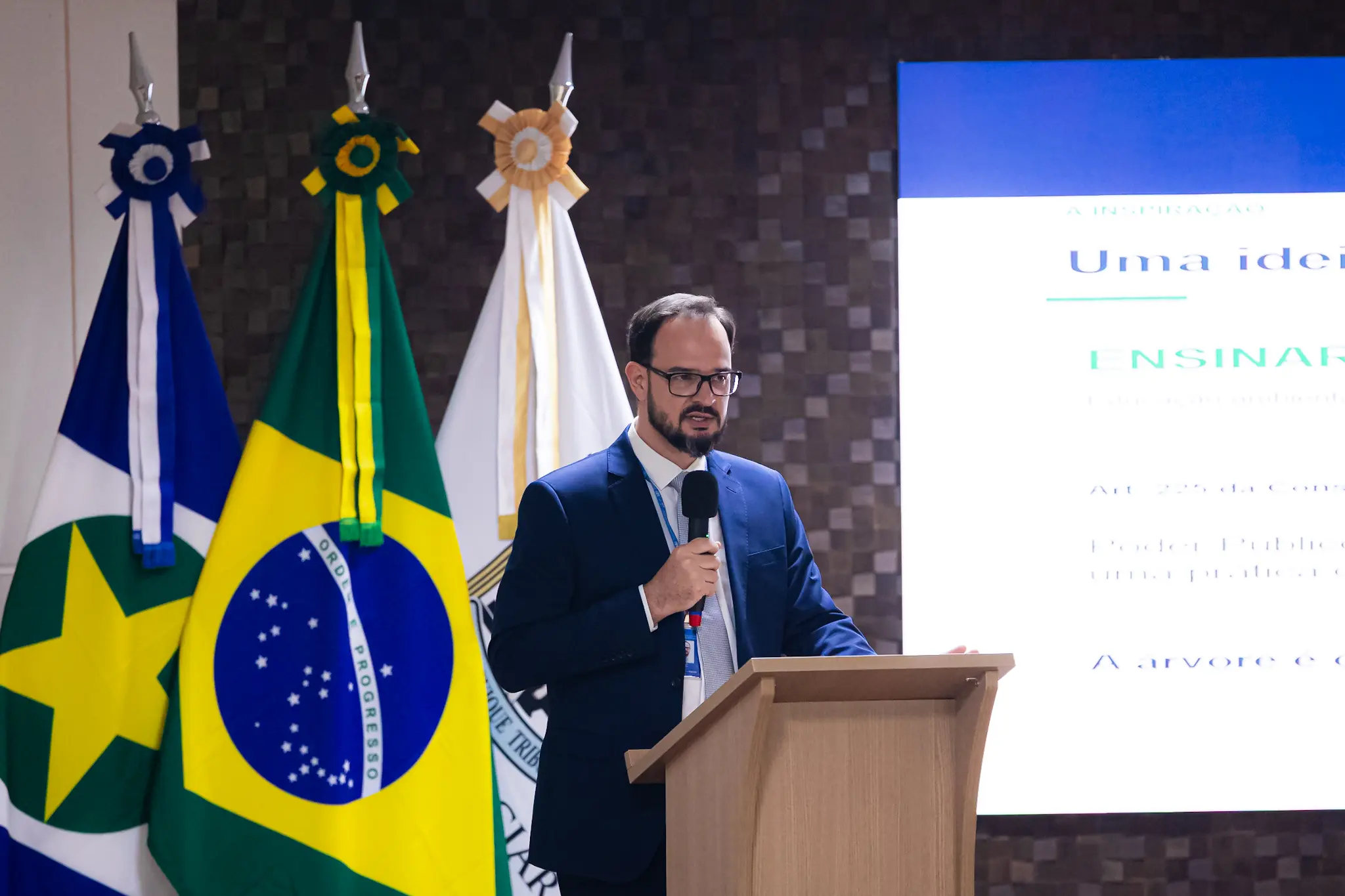 Homem de óculos, barba e terno azul-marinho, discursa ao microfone em púlpito com telão de apresentação e bandeiras ao fundo.