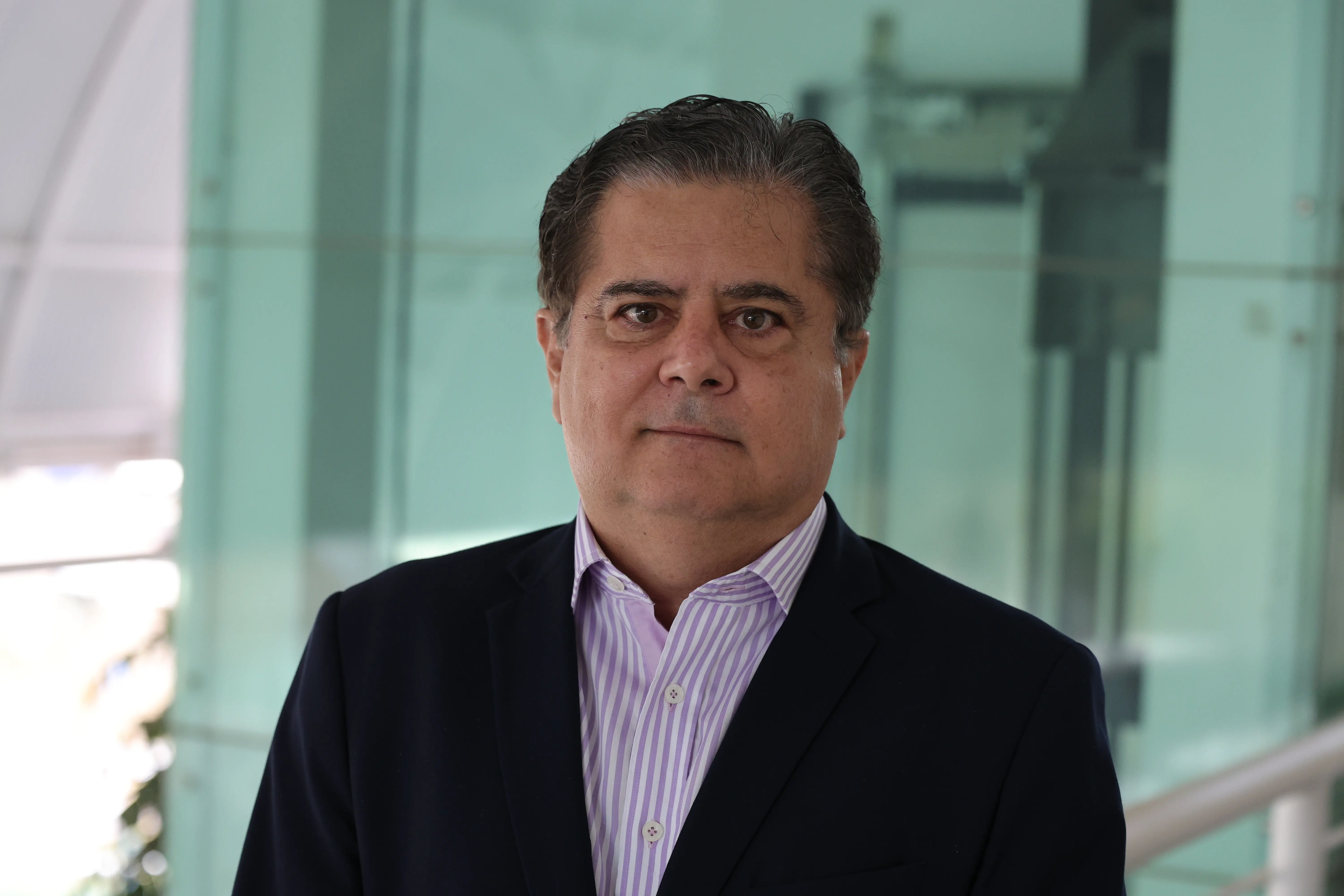 Retrato de um homem de cabelos escuros, de óculos, vestindo paletó escuro sobre camisa de listras roxas e brancas, posicionado em frente a uma parede de vidro.