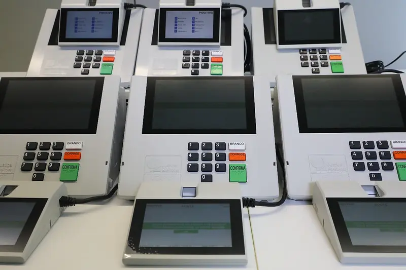 Diversas urnas eletrônicas brasileiras organizadas em fileiras sobre uma mesa clara. Na parte frontal, estão conectados os terminais de mesário com tela digital acesa.