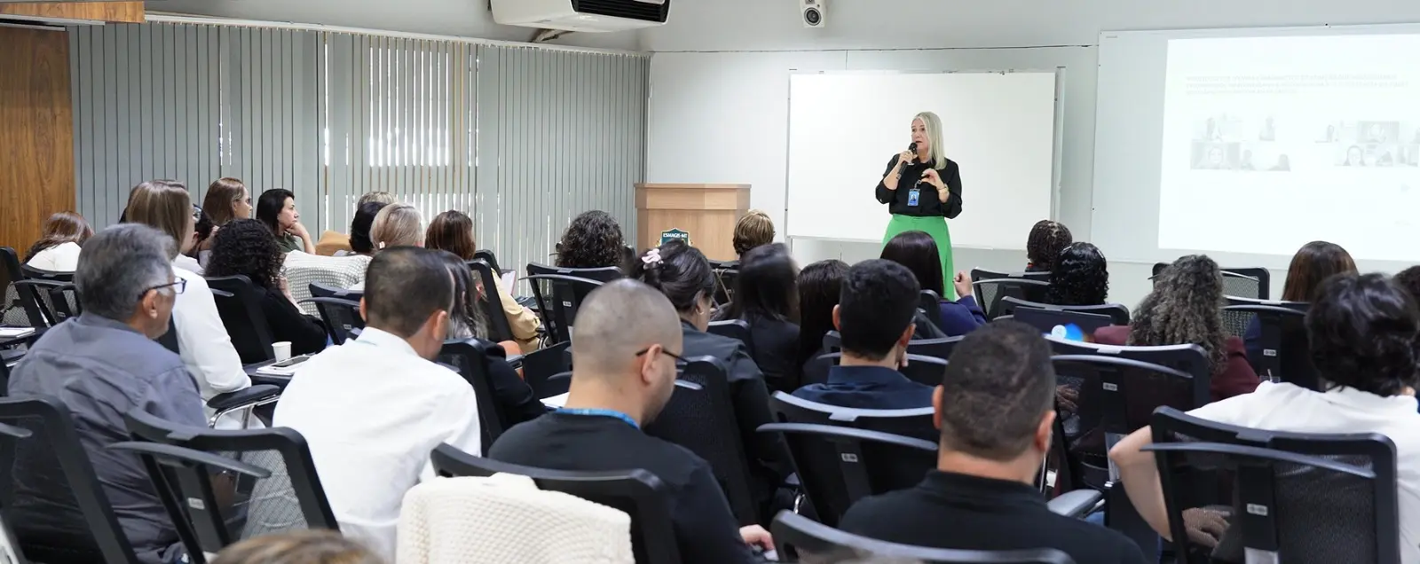 Foto horizontal que mostra uma sala de aula cheia de pessoas sentadas nas carteiras. à frente, uma mulher branca e loira fala ao microfone.