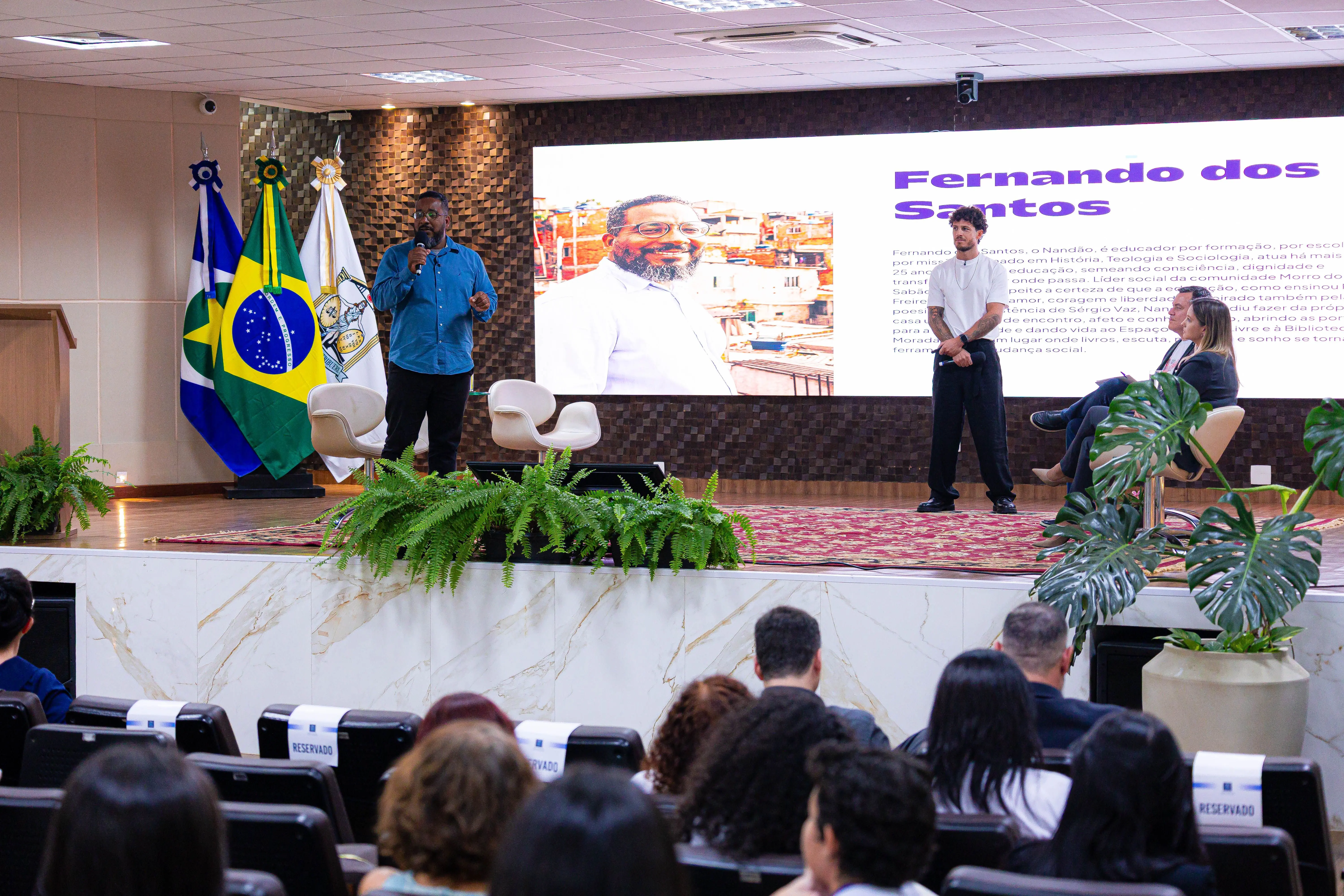 Homem de camisa azul fala ao microfone durante palestra do projeto Boyzinho de Respeito, enquanto outro participante e uma mediadora acompanham no palco.
