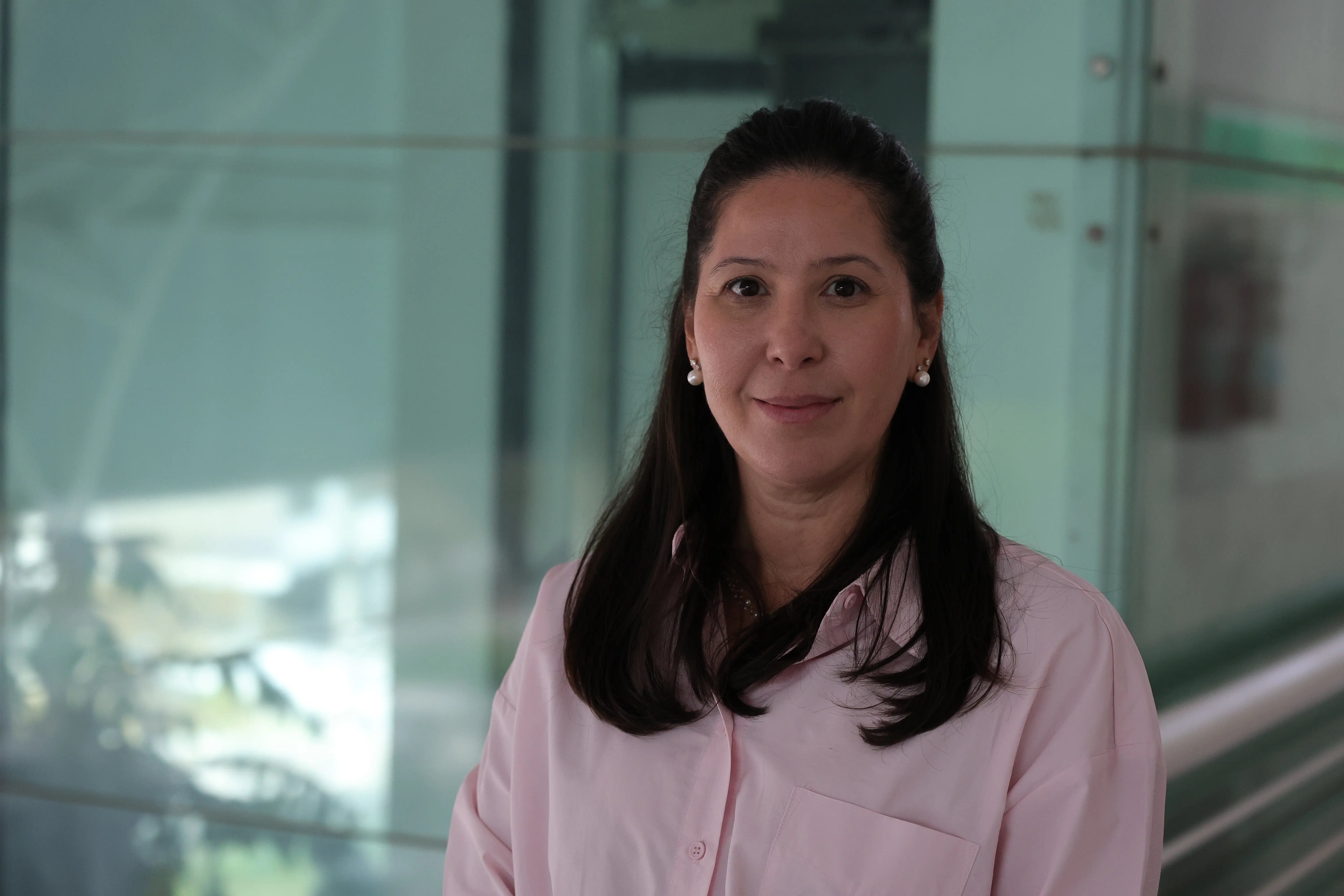 Retrato de uma mulher de cabelos escuros longos e camisa rosa clara. Ela está posicionada no centro da imagem em um ambiente interno com divisórias de vidro ao fundo.