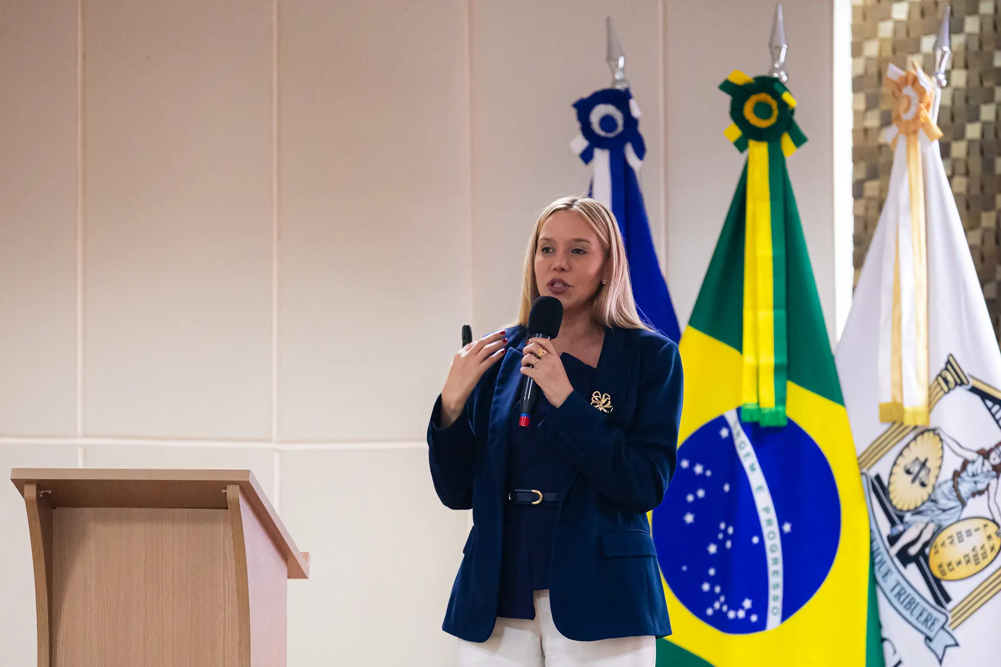 Mulher loira de blazer azul-marinho e calça branca, fala ao microfone diante das bandeiras de MT, Brasil e do TJMT.