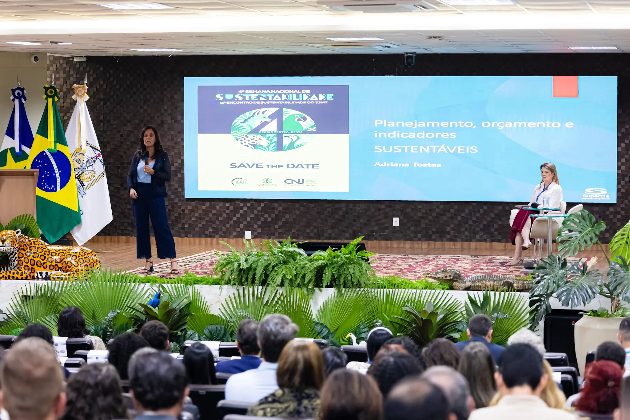 Mulher palestra no palco diante de telão com o tema "Planejamento, orçamento e indicadores SUSTENTÁVEIS". No palco, há bandeiras, folhagens e esculturas de animais. Platéia assiste em primeiro plano.