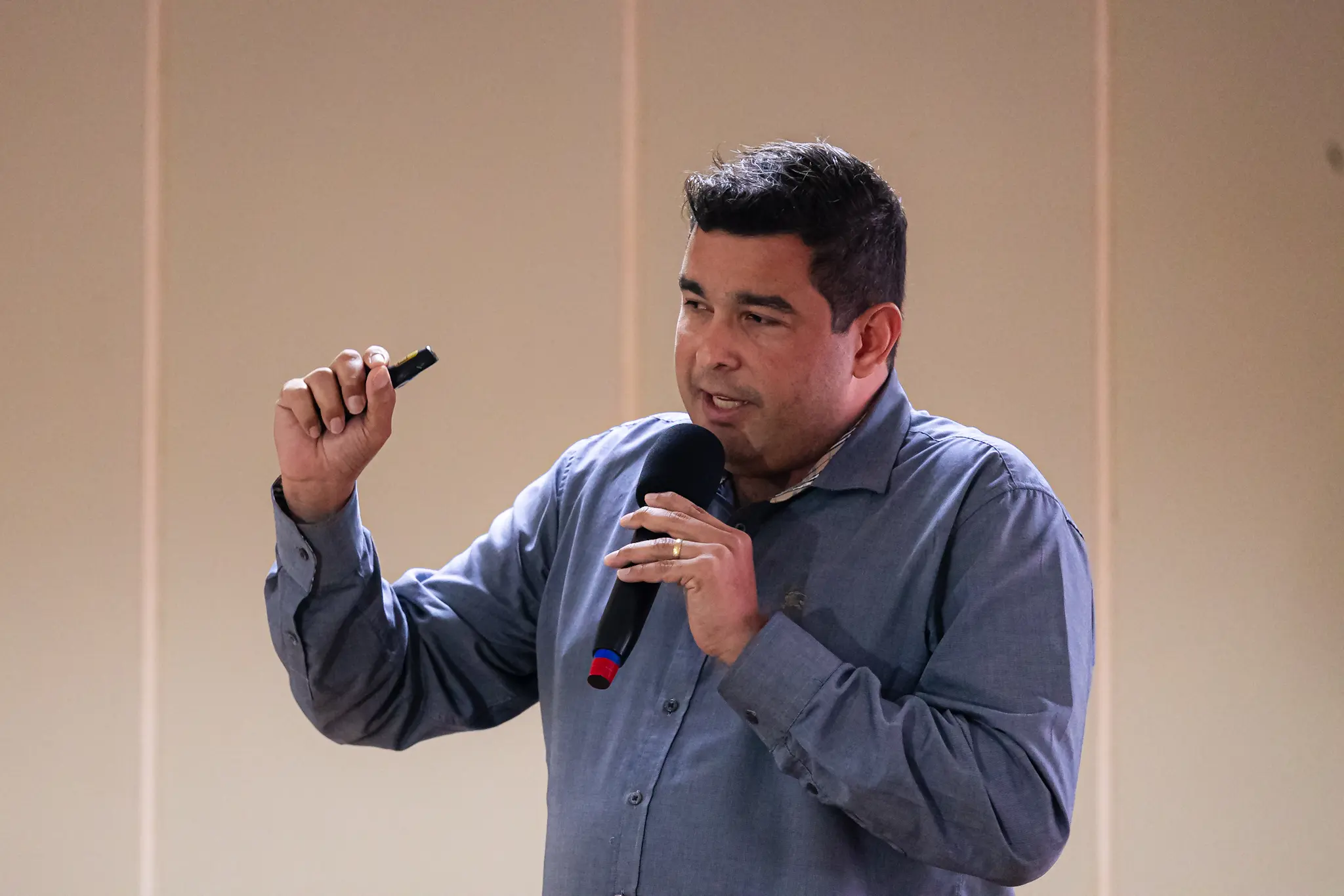 Homem de cabelos escuros, camisa azul, fala ao microfone gesticulando com a outra mão, fundo bege neutro.