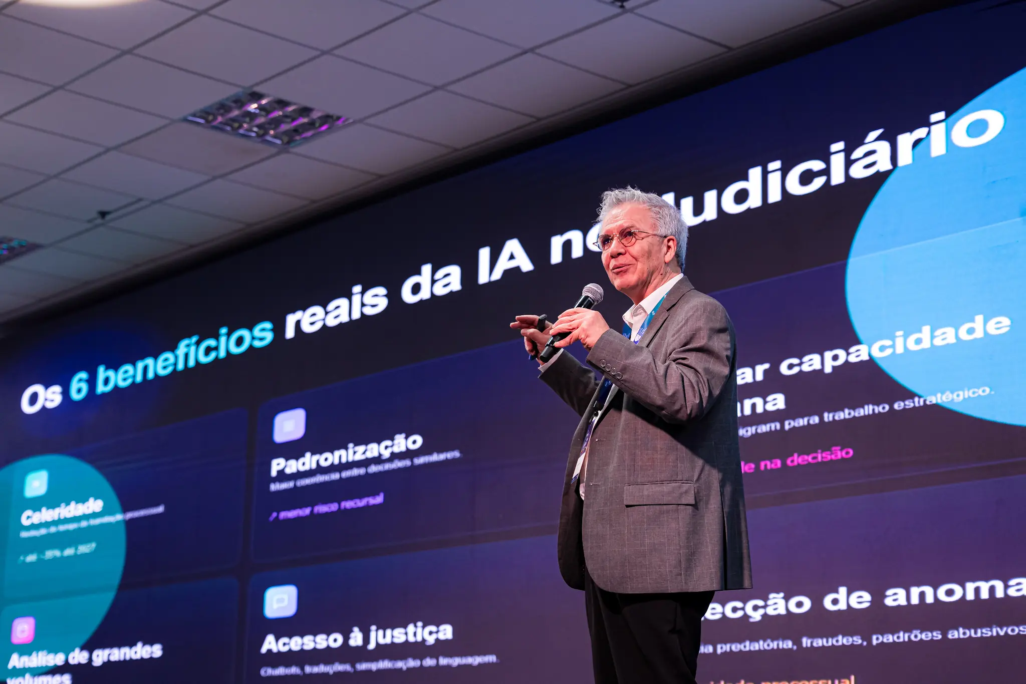 Homem de cabelos brancos, óculos e paletó cinza fala ao microfone em um palco. Ao fundo, um telão escuro apresenta o título "Os 6 benefícios reais da IA no Judiciário" acompanhado de quadros explicativos com tópicos como "Celeridade" e "Padronização".