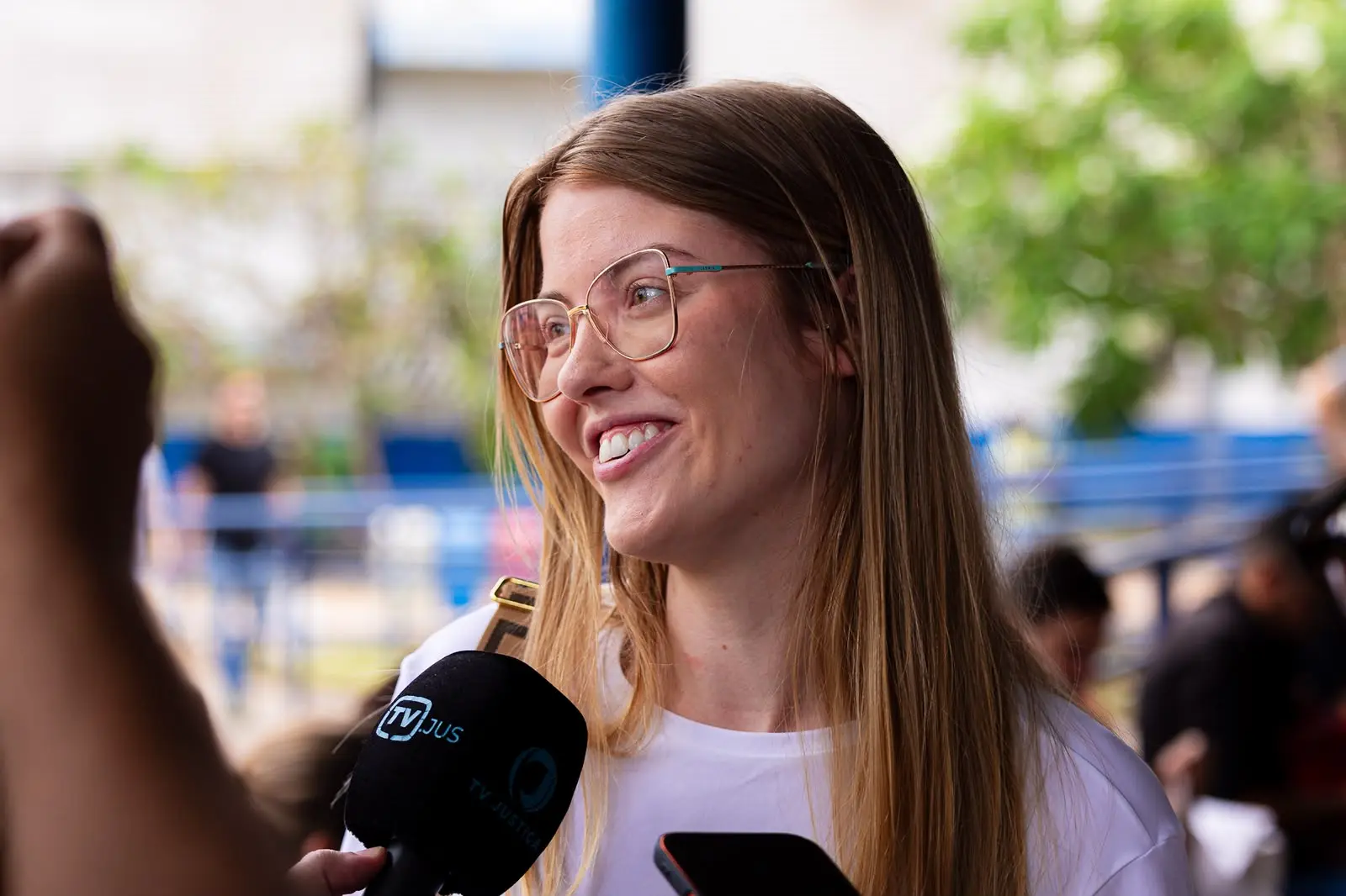 Plano médio de uma mulher jovem de pele clara, cabelos loiros lisos e óculos de grau finos, sorrindo levemente enquanto fala. À sua frente, há um microfone preto com o logotipo "TV JUS". Ao fundo, ambiente externo desfocado com tons azuis e verdes.
