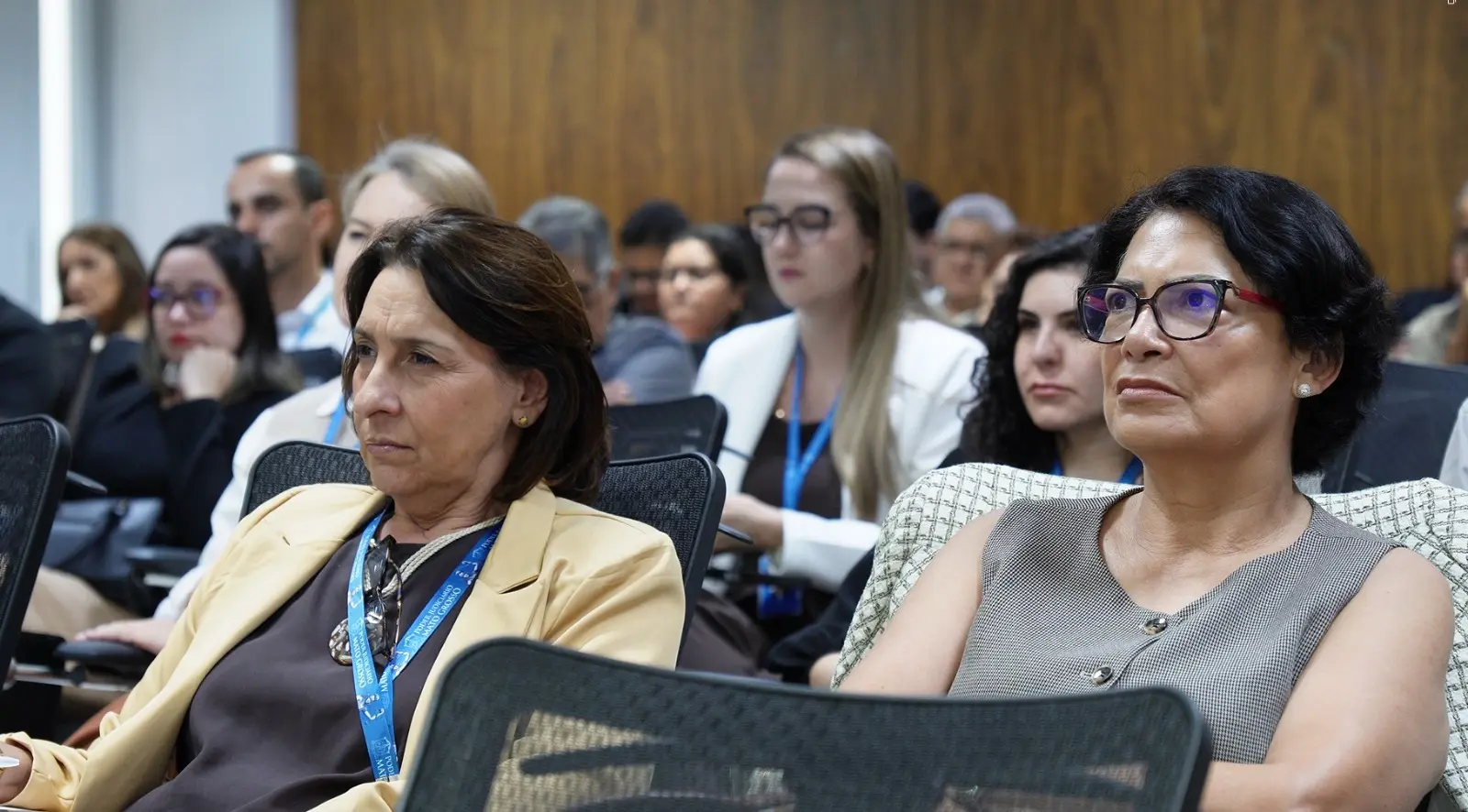 Foto horizontal que mostra o público do Encontro Anual de Gestores. São várias pessoas sentadas em carteiras, na sala de aula, olhando para a frente. Em primeiro plano, aparecem duas mulheres.