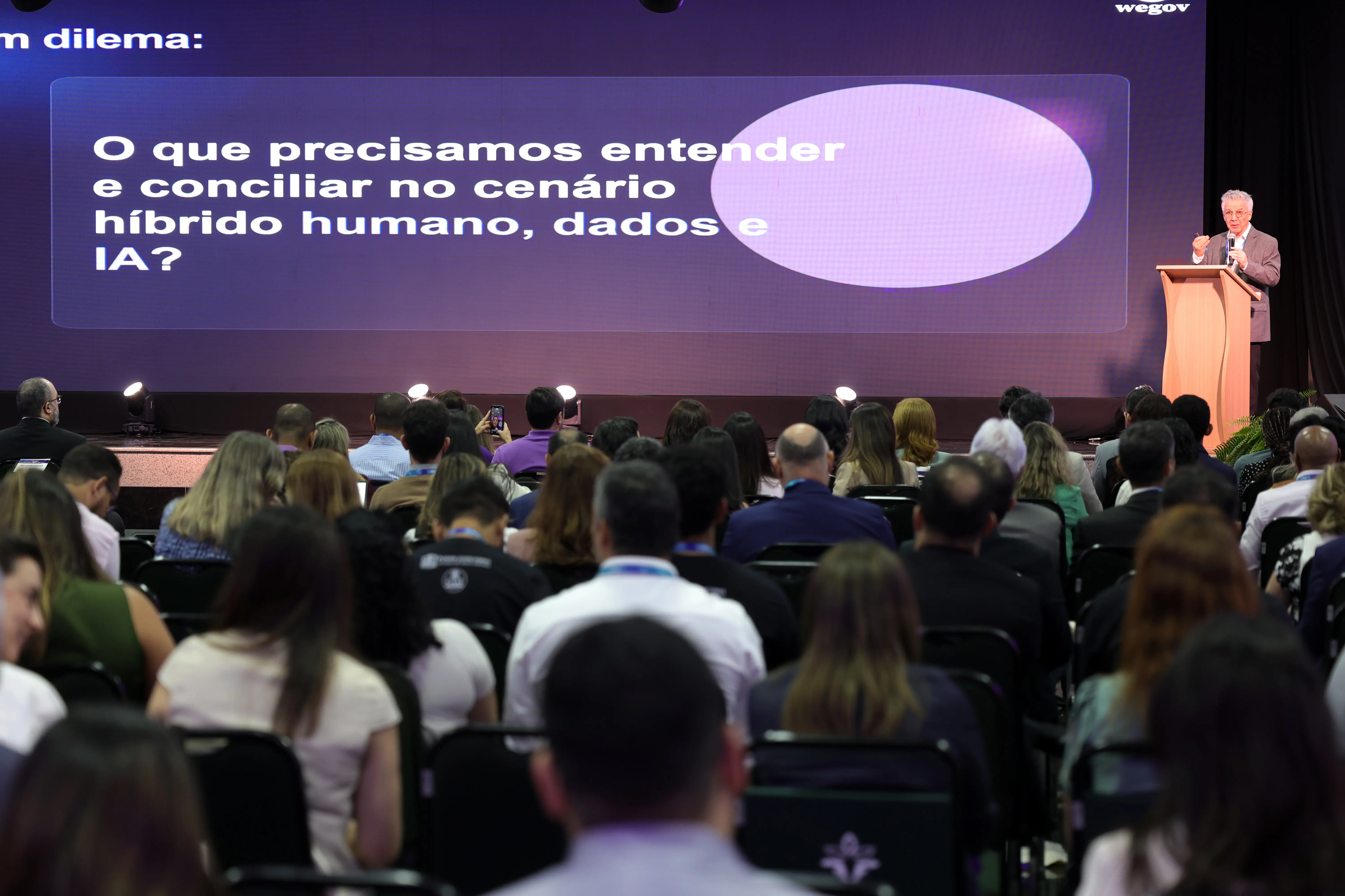 Visão posterior de auditório lotado durante palestra. Na frente, à direita, um homem de cabelos brancos e paletó discursa em um púlpito. O telão exibe o texto: "O que precisamos entender e conciliar no cenário híbrido humano, dados e IA?".