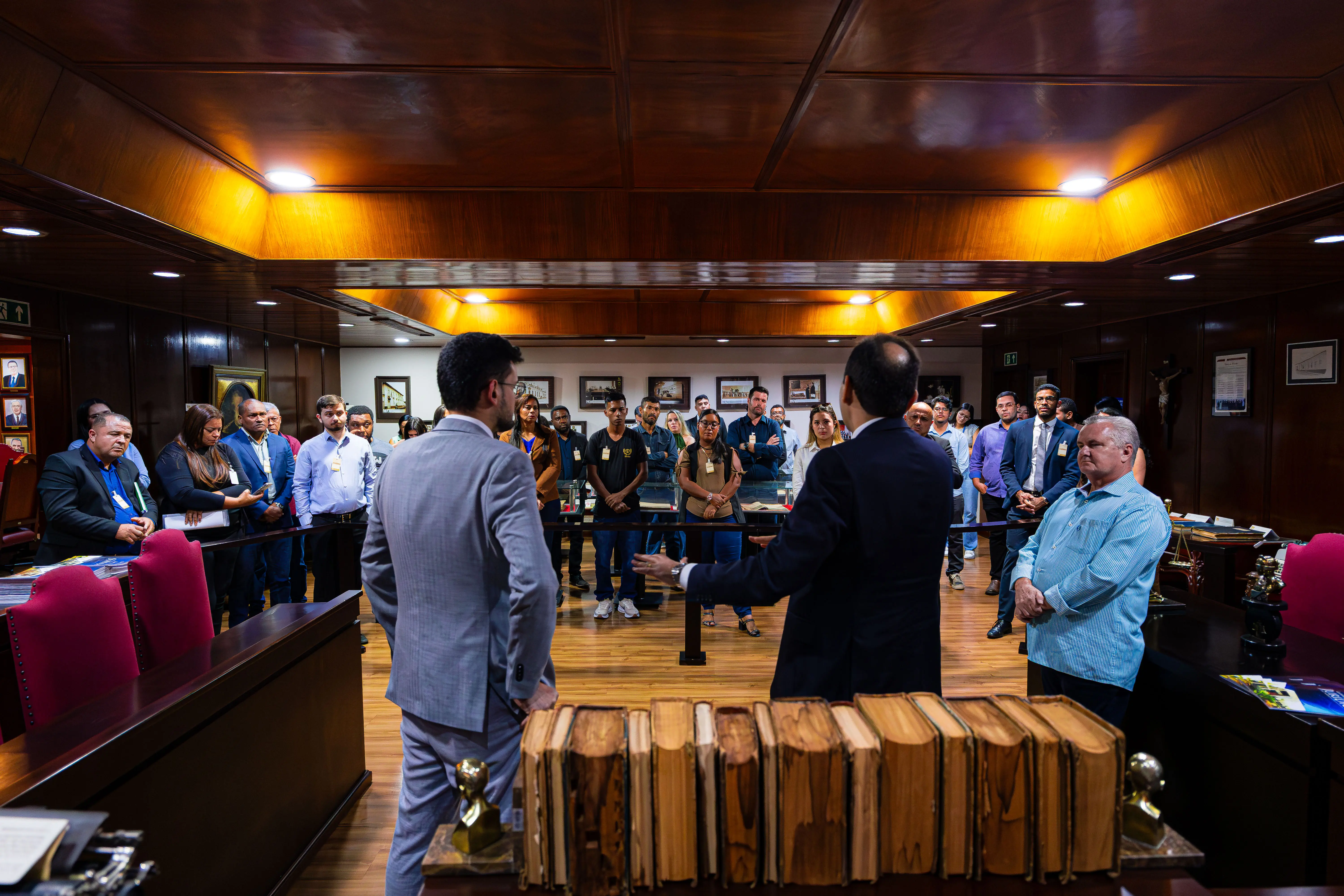 Grupo de participantes reunidos no Memorial do Judiciário observa explicação de dois homens de terno, cercados por livros antigos, retratos na parede e mobiliário de madeira.