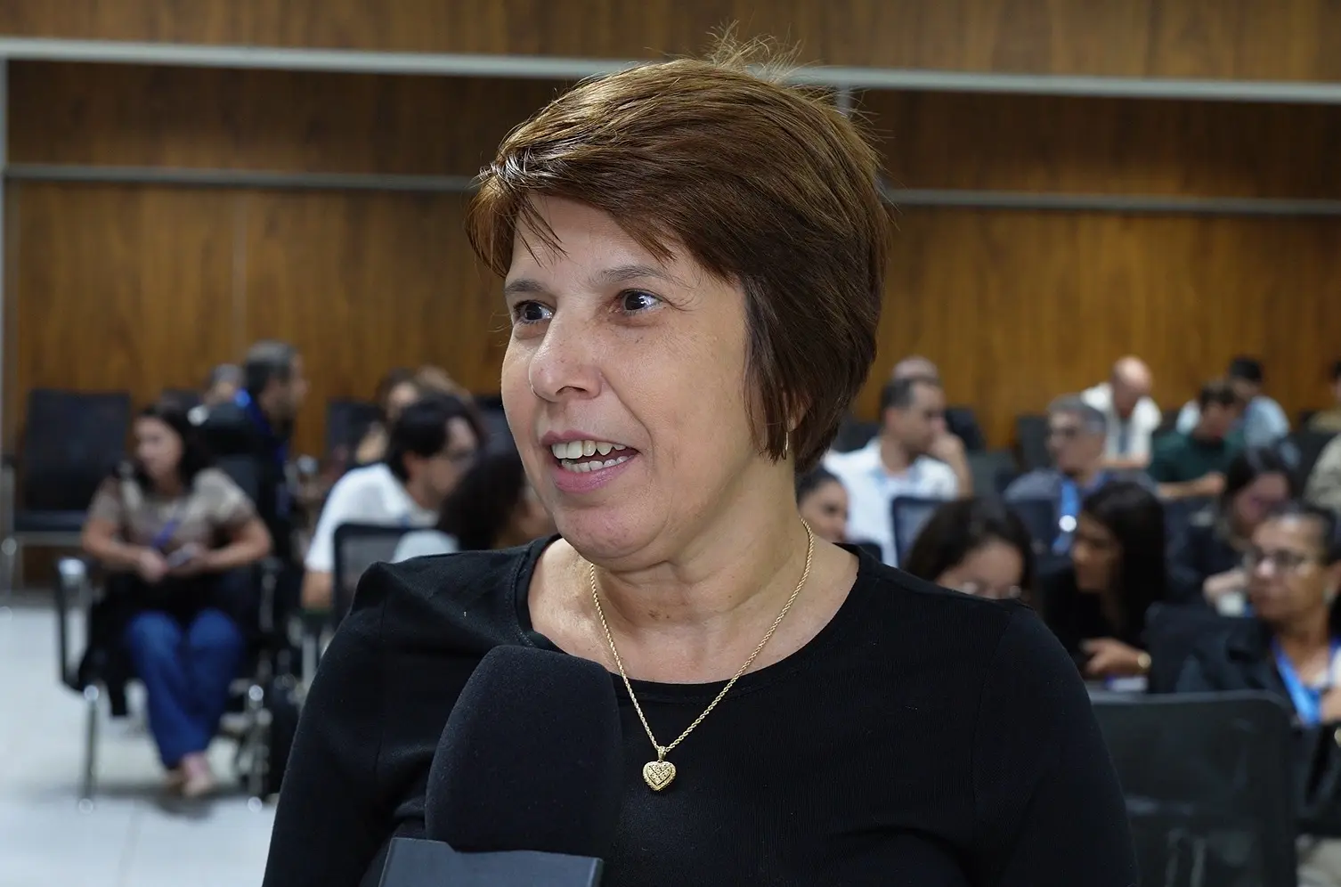 Foto horizontal que mostra a gestora geral do Fórum de Barra do Garças, Elizângela Schweig, durante entrevista à TV TJMT. Ela é uma mulher de pele clara, olhos castanhos, cabelos curtos, lisos e castanhos, com blusa preta e colar com pingente de coração dourado. Ao fundo, ambiente de sala de aula.