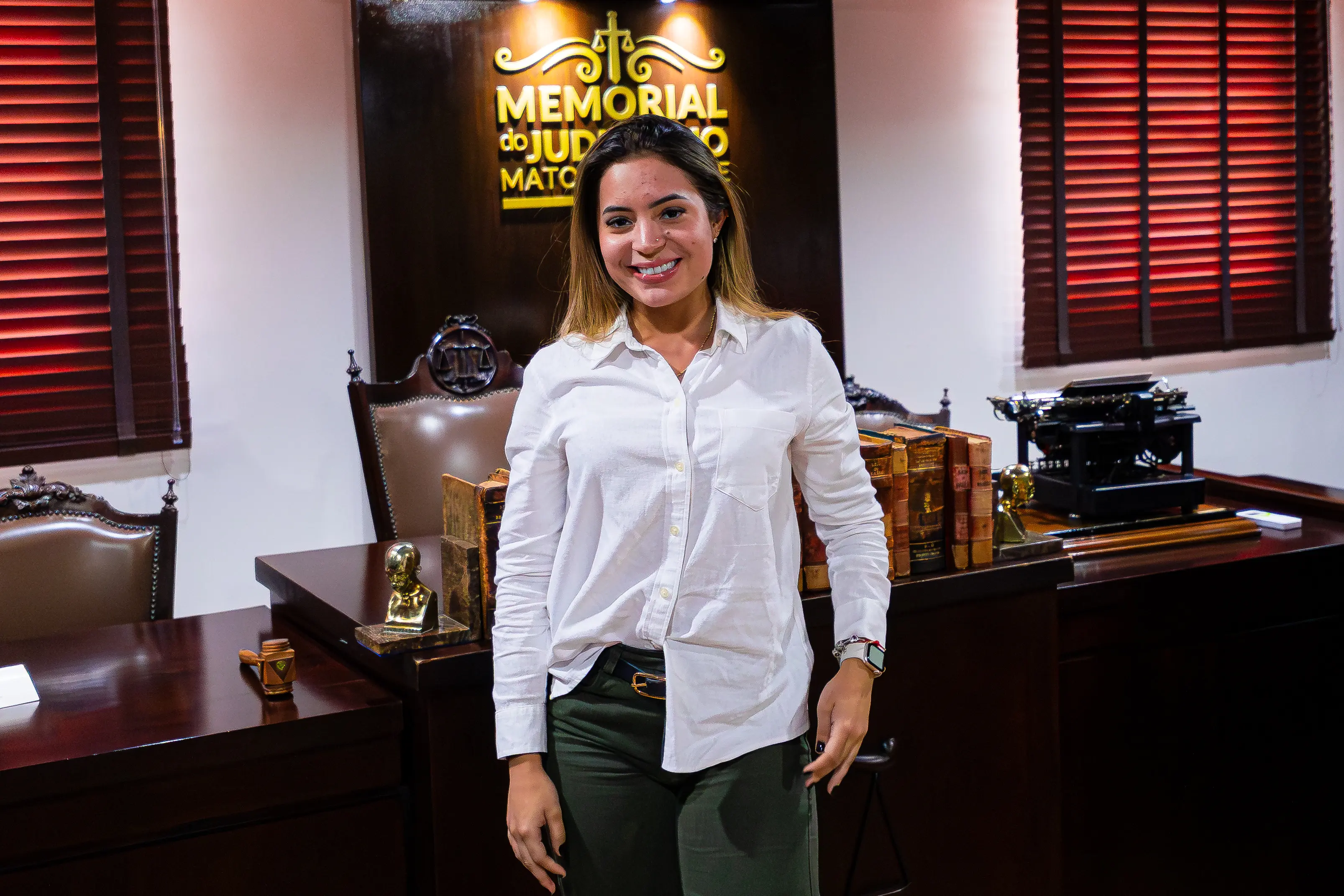 Mulher de cabelos castanhos, sorridente, veste camisa branca e calça verde, posa no Memorial do Judiciário de Mato Grosso, com estantes de livros antigos e máquina de escrever ao fundo.