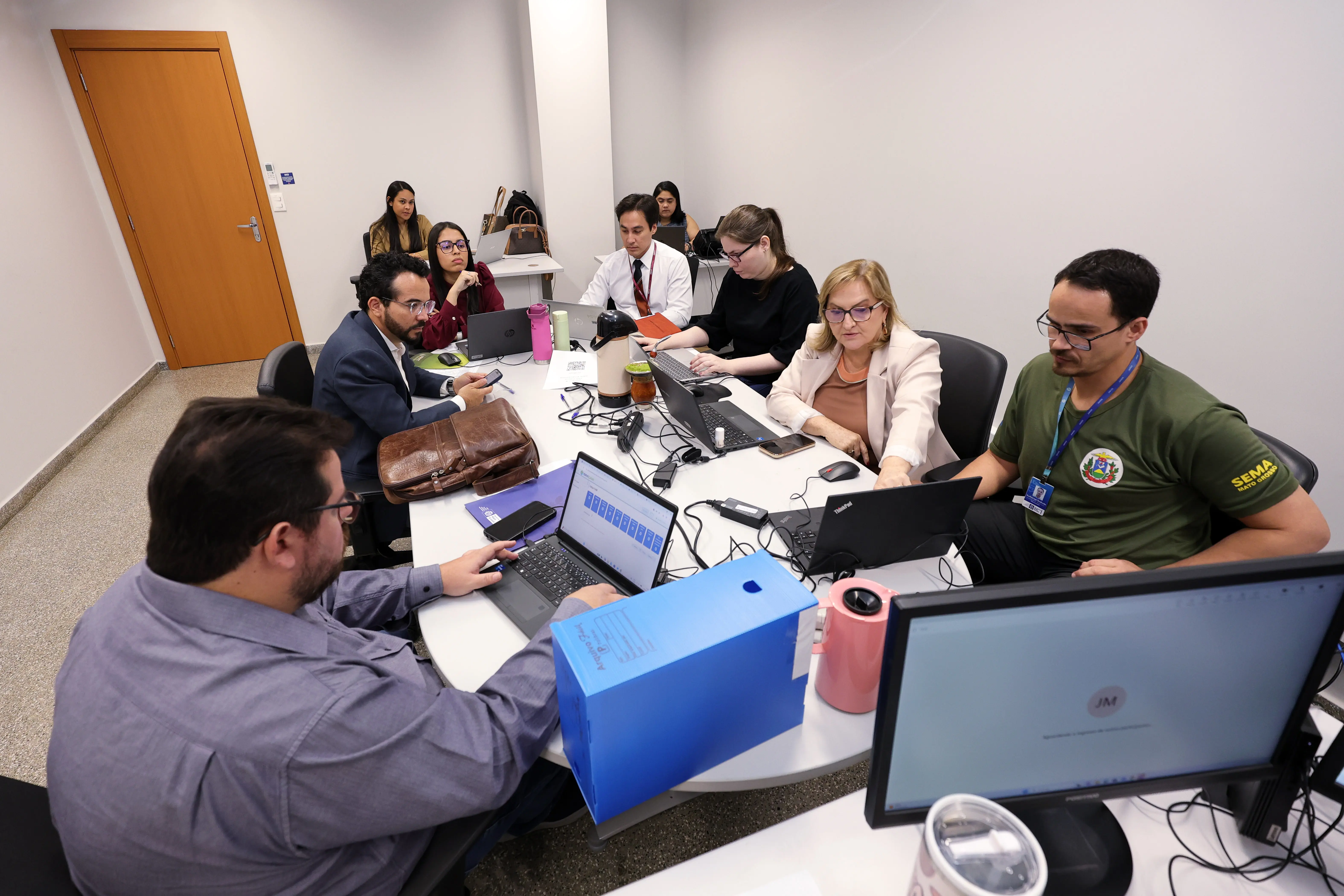 Visão em ângulo amplo de uma sala com seis pessoas reunidas ao redor de uma mesa oval de trabalho cheia de notebooks, cabos, uma garrafa térmica e uma caixa de arquivo azul. Um homem em primeiro plano à esquerda digita em um notebook. Ao fundo, duas pessoas trabalham em bancadas separadas.