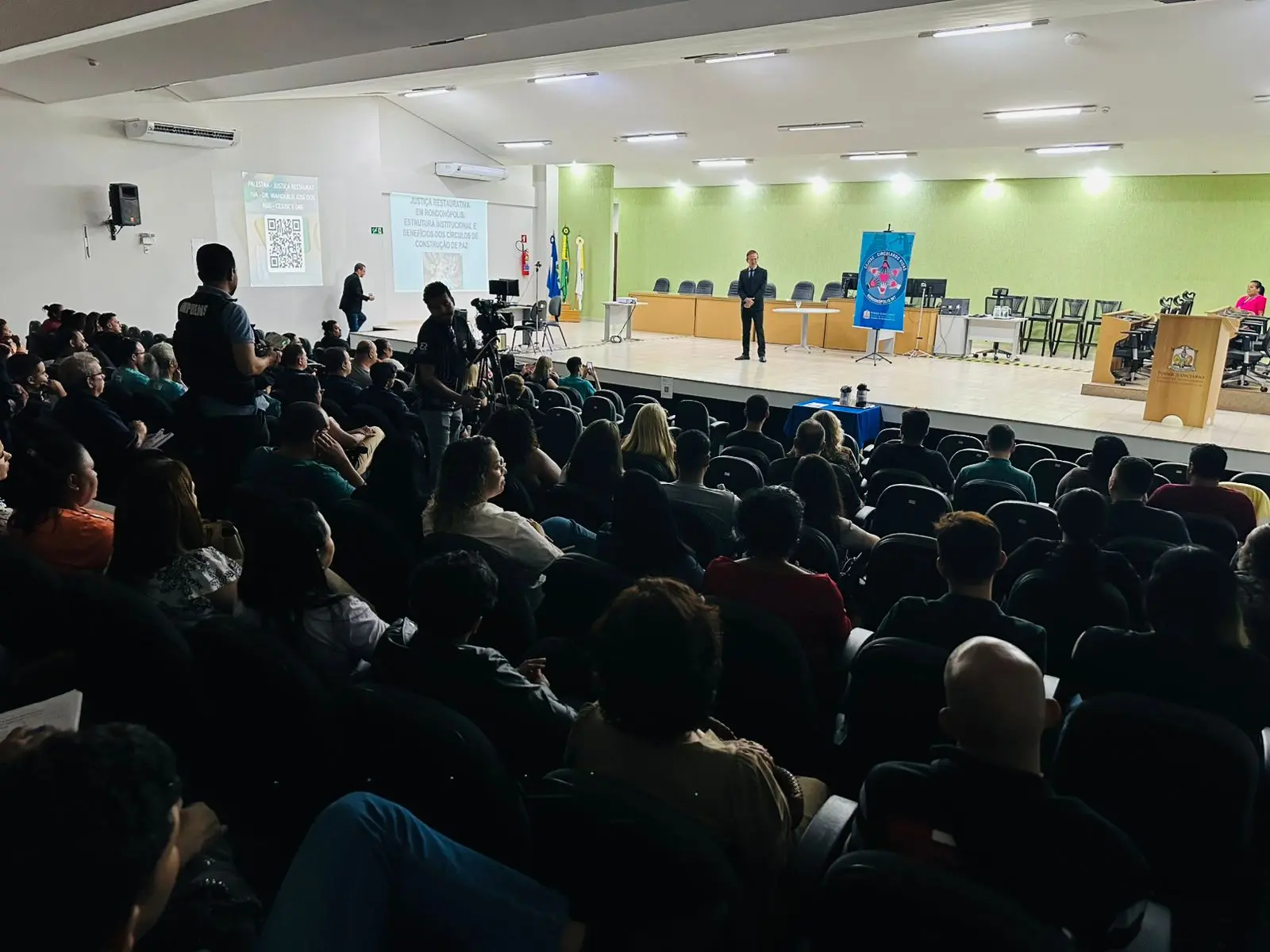 Auditório cheio registra evento institucional. Juiz ao centro do palco, banner do Cejusc à direita e bandeiras ao fundo. Público ocupa quase todas as poltronas.