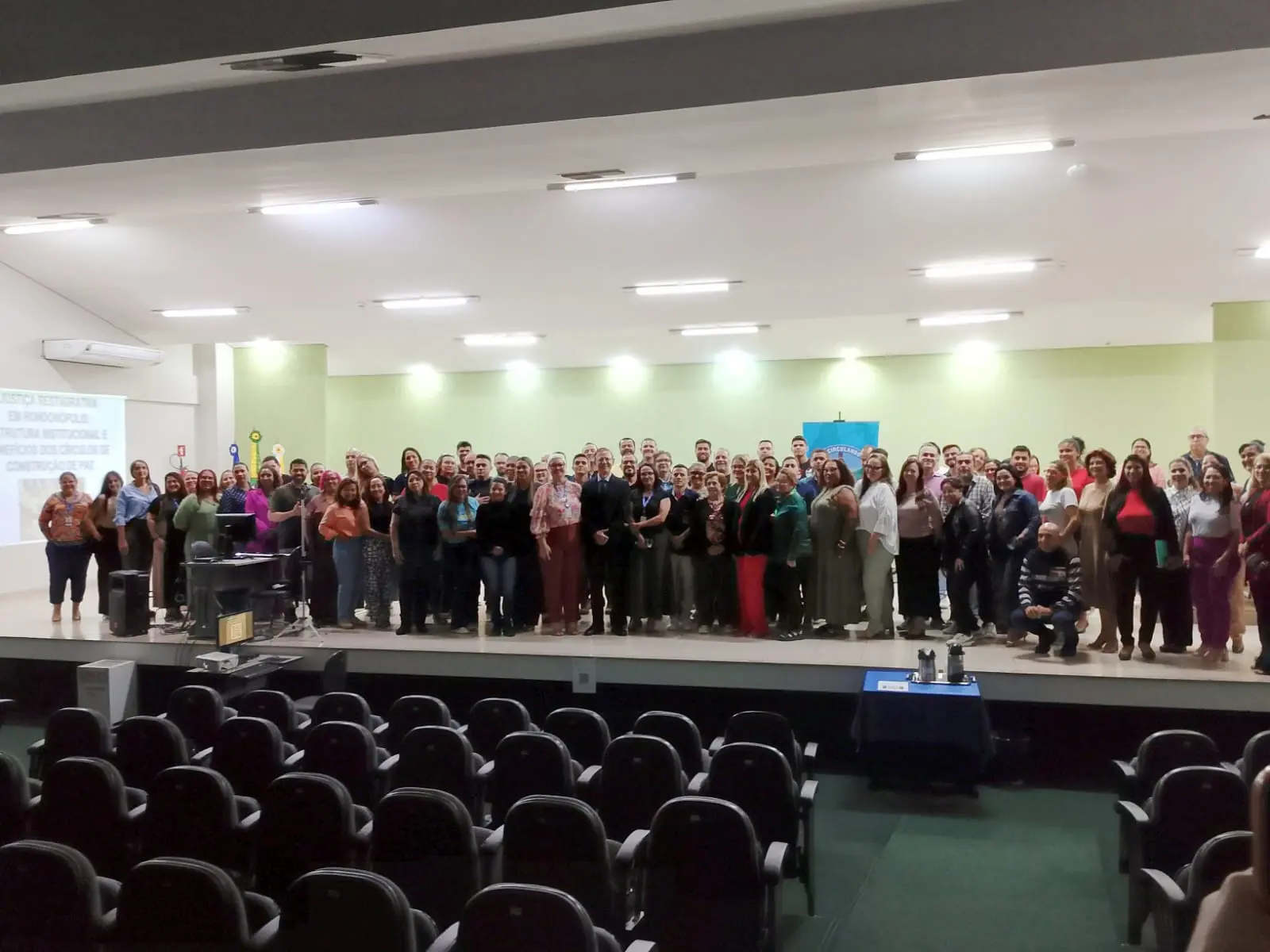 Grande grupo posa no palco após o evento. Participantes reunidos lado a lado para foto oficial. Auditório ao fundo com cadeiras vazias na parte frontal.