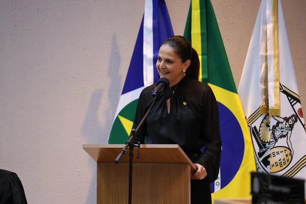 Foto horizontal em plano médio que mostra a presidente da OAB-MT, Gisela Alves Cardoso, falando ao microfone, na tribuna do plenário do TJMT. Ela é uma mulher branca, magra, de cabelos castanhos presos, usando calça preta e blusa de manga comprida preta. 