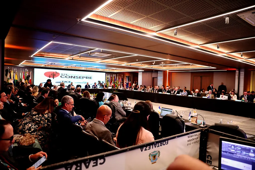 Amplo auditório com dezenas de pessoas sentadas em mesas longas dispostas em 