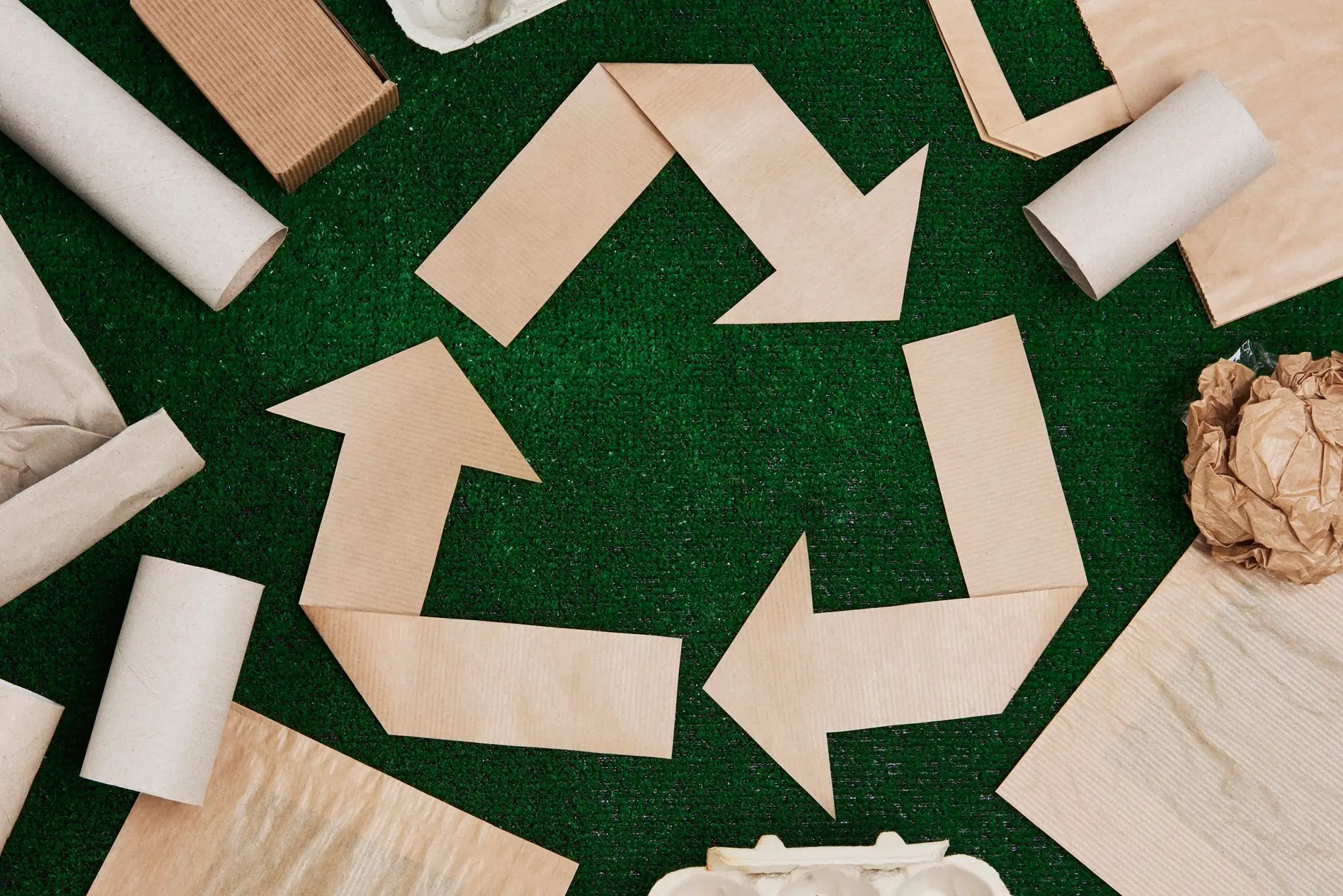 Setas de papelão formam símbolo da reciclagem sobre fundo verde, cercadas por rolos e embalagens de papel. A imagem representa reaproveitamento, economia circular e práticas sustentáveis.
