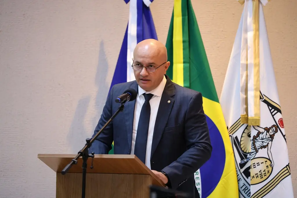Foto horizontal em plano médio que mostra o primeiro subdefensor público-geral, Rogério Borges Freitas, falando ao microfone, na tribuna do plenário do TJMT. Ele é um home branco, careca, usando camisa branca, gravata e terno azul marinho e óculos de grau. 