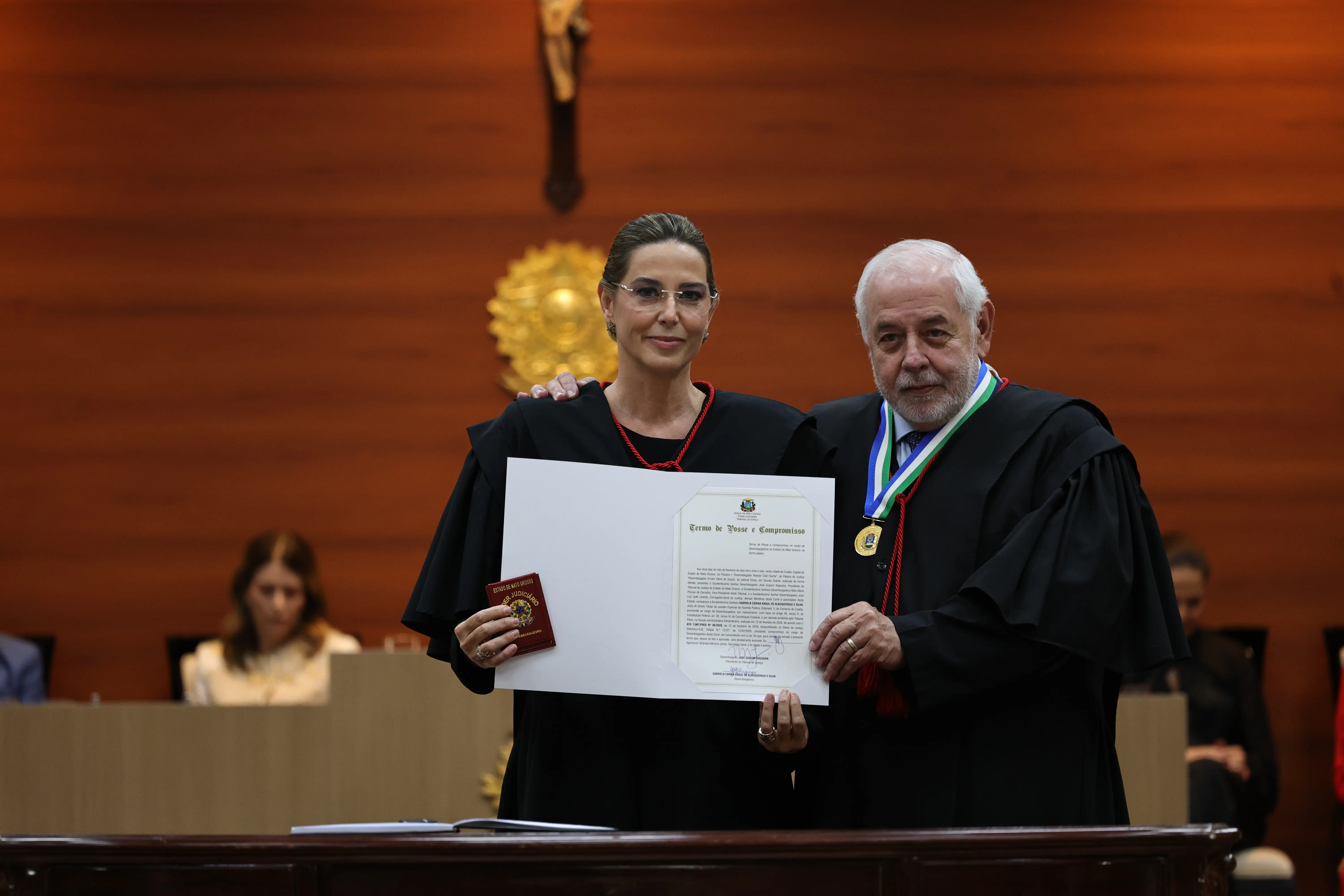 Mulher e homem idoso em trajes de magistrados posam lado a lado segurando certificado. Ele usa medalha com cordão verde, branco e azul no pescoço. Ao fundo, parede de madeira com crucifixo dourado.