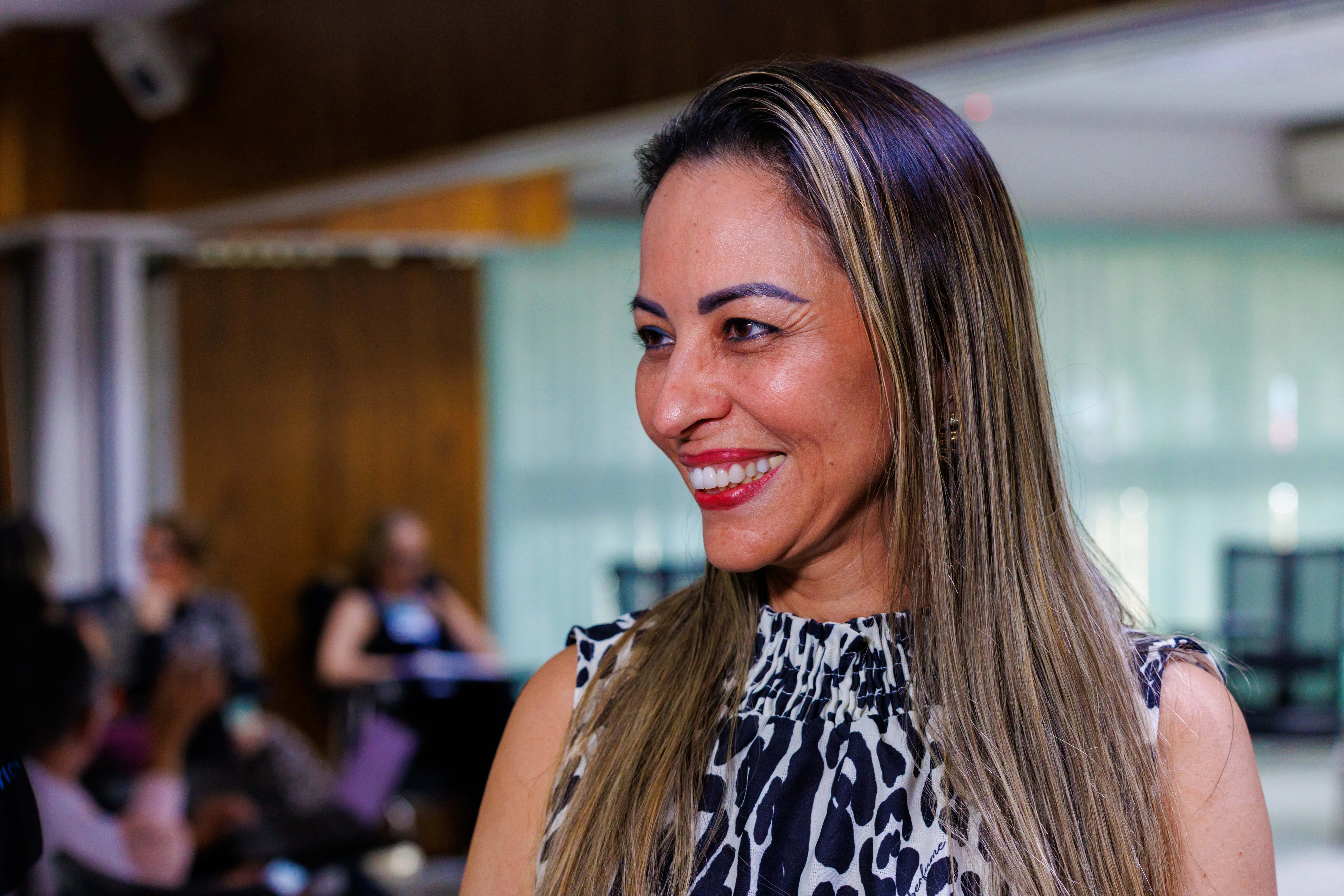 Rogéria Borges é uma mulher de cabelos longos e lisos sorri em ambiente interno iluminado. Ela veste blusa estampada em preto e branco. Ao fundo, pessoas desfocadas participam de atividade em sala ampla com janelas de vidro.
