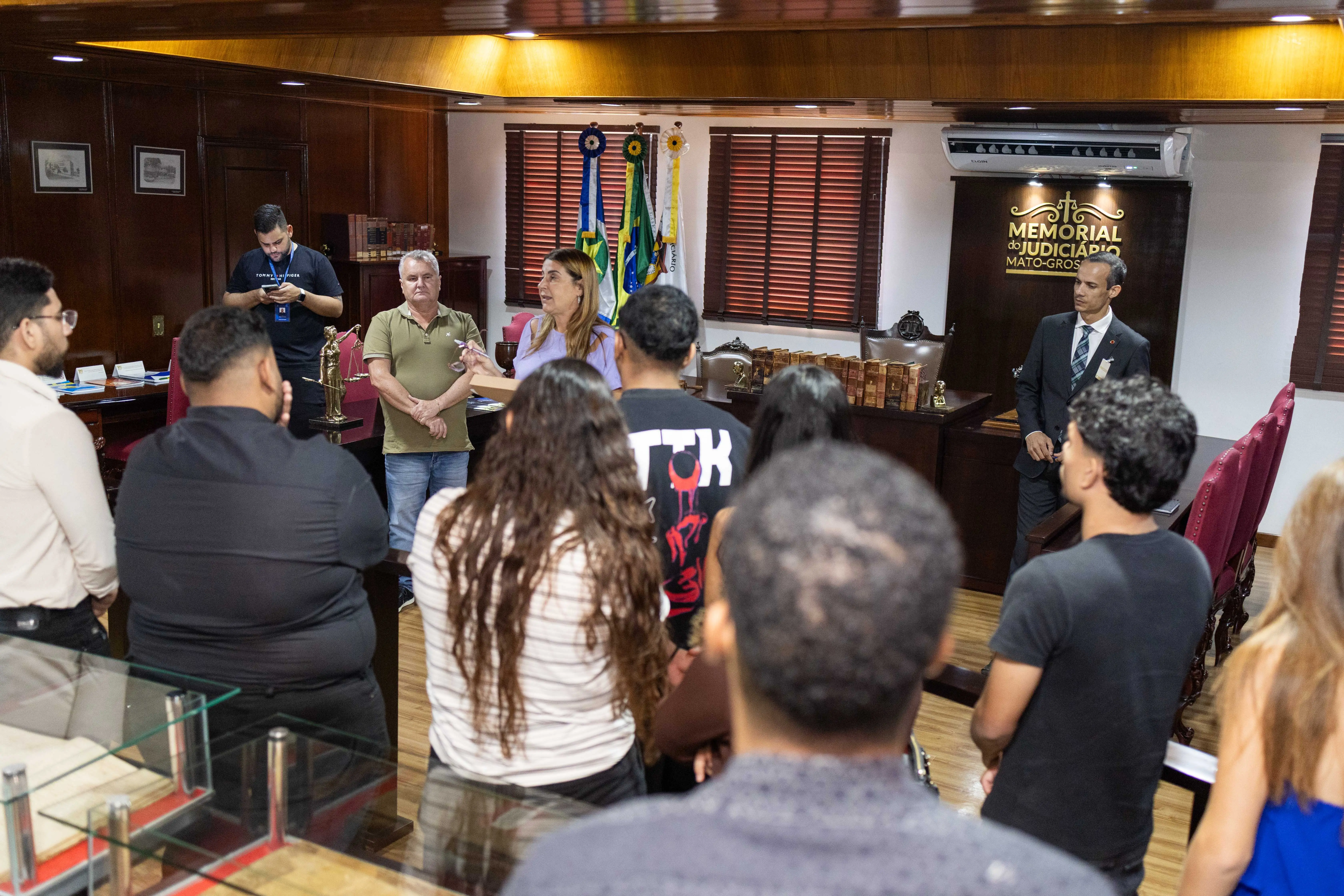 Grupo de pessoas visita sala do Memorial do Judiciário de Mato Grosso. Uma mulher fala ao centro, enquanto outras pessoas escutam atentas. Ao fundo, há bandeiras, livros antigos e móveis em madeira escura.