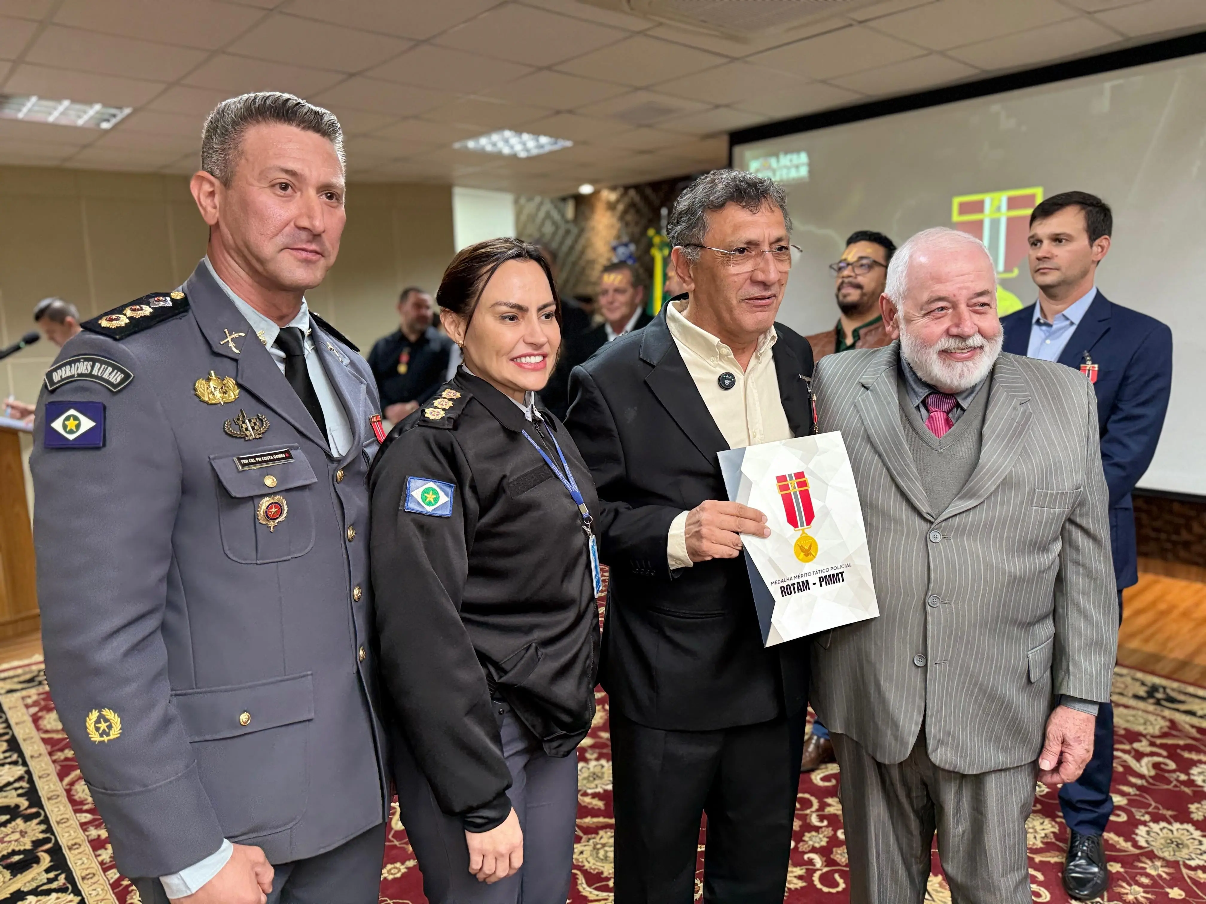 Grupo de autoridades e homenageados posam sorridentes. Um dos homens segura certificado com brasão da ROTAM. Estão próximos ao telão com projeção da medalha. O ambiente é decorado com tapete vermelho