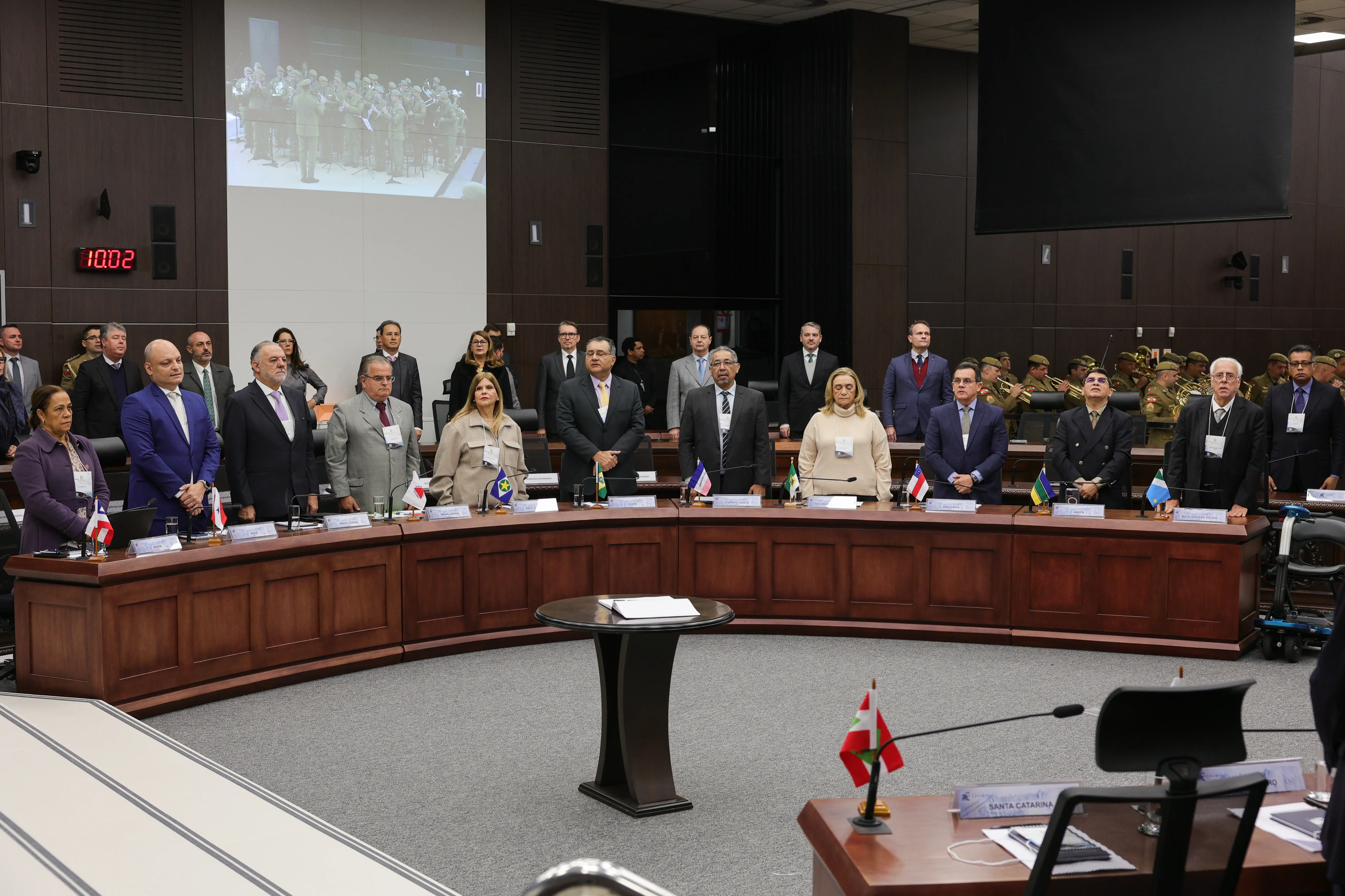 Imagem mostra uma grande reunião em auditório. Pessoas de terno e trajes formais estão em pé, dispostas em semicírculo ao redor de uma mesa de madeira. Ao fundo, há plateia sentada e telão exibindo uma mulher falando.