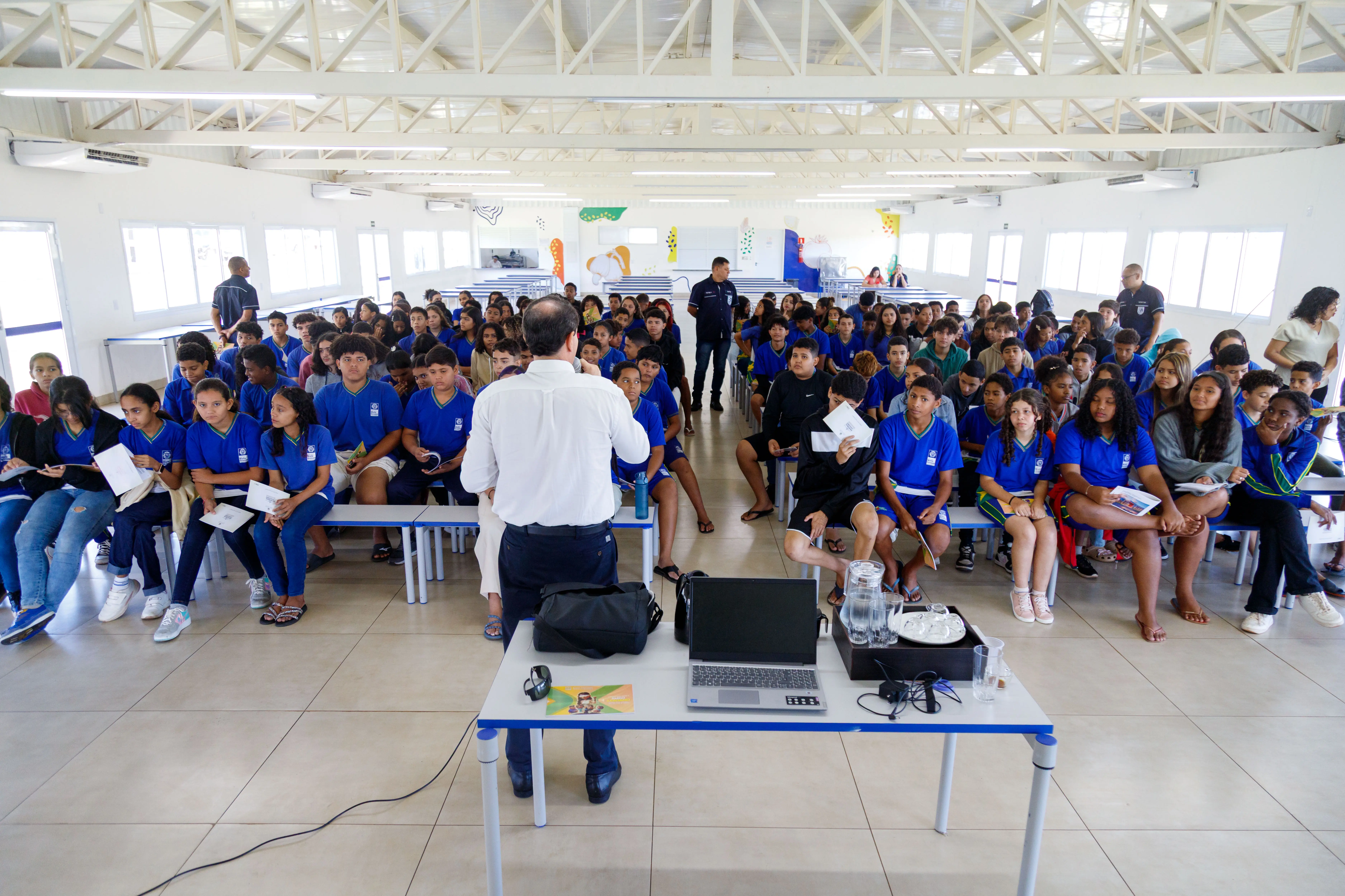 A imagem mostra um auditório amplo com um grande grupo de estudantes sentados em bancos, a maioria vestindo camisetas azuis. Um homem de camisa branca está de costas para a câmera, apresentando uma palestra
