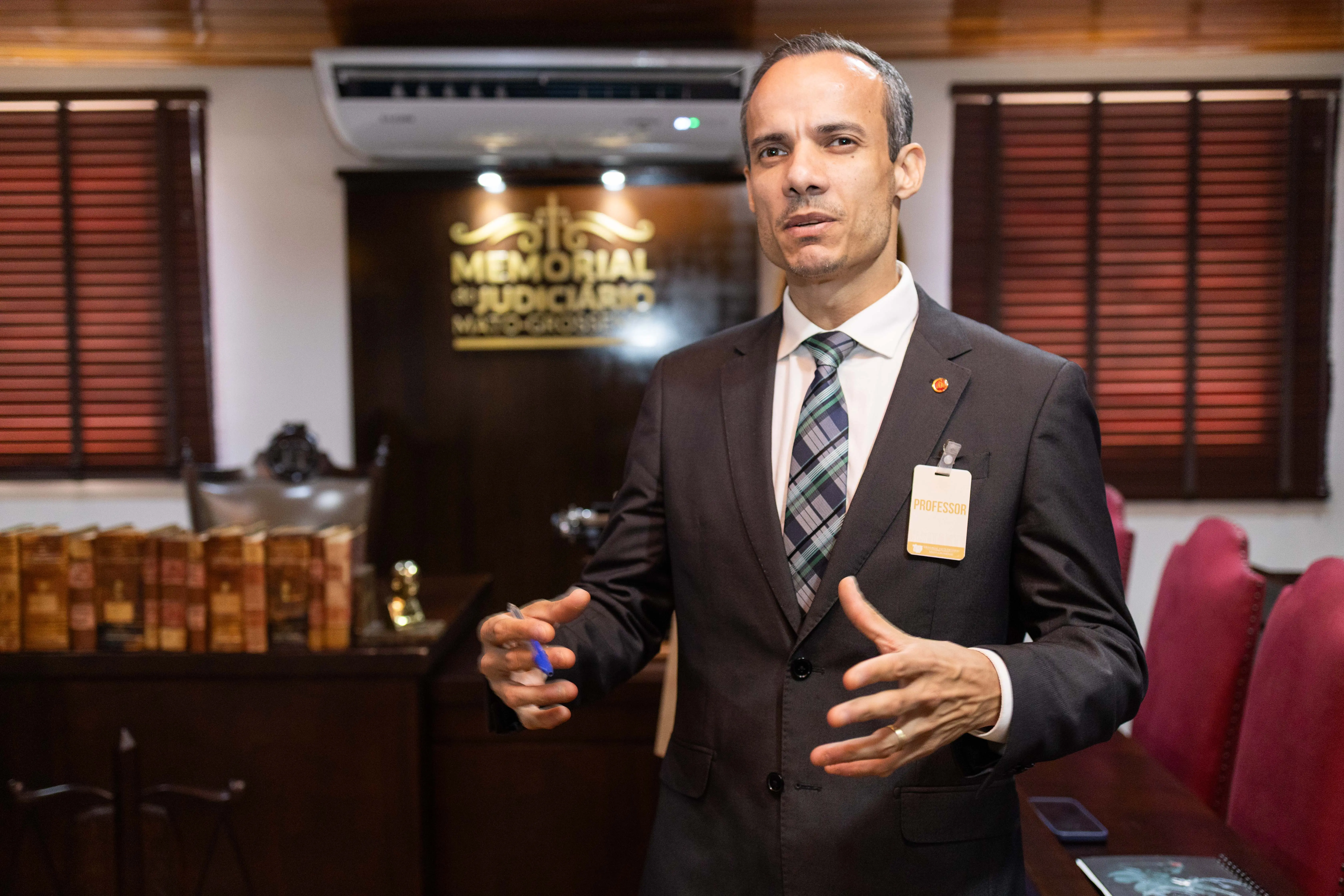 O professor Thomas Ubirajara, vestido com um terno escuro, camisa branca e gravata listrada, está em pé gesticulando com as mãos. Ele usa um crachá de "Professor" no bolso do paletó. Ao fundo, um letreiro dourado indica "Memorial do Judiciário Mato-Grossense" e há estantes com livros antigos.