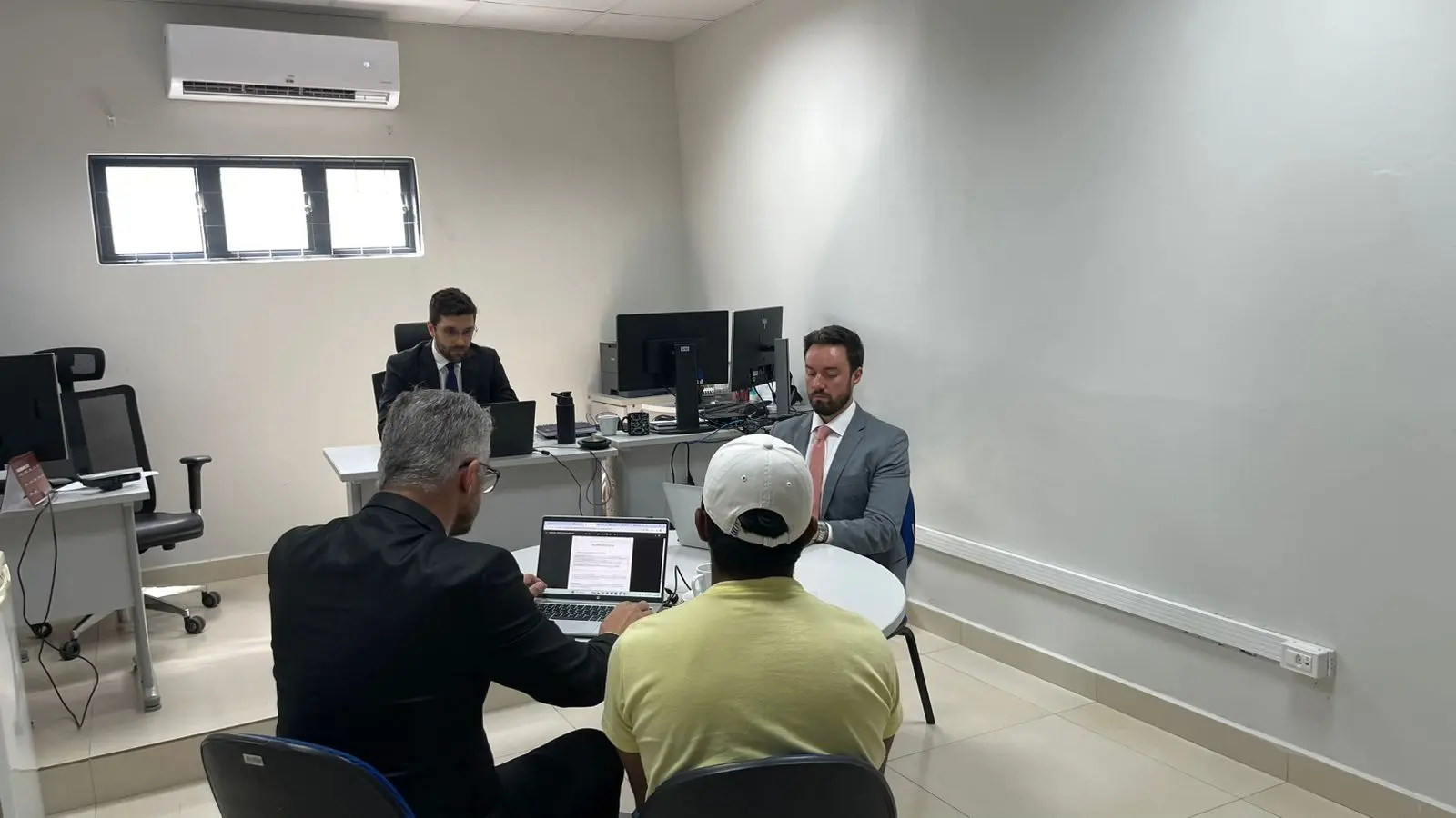 Sala com duas mesas e computadores. Dois homens de terno estão sentados em posições separadas; um deles está à frente de uma mesa redonda, na qual outras duas pessoas (de costas, uma de terno e outra de camiseta amarela com boné) participam da reunião. Ao fundo o magistrado realiza audiência.