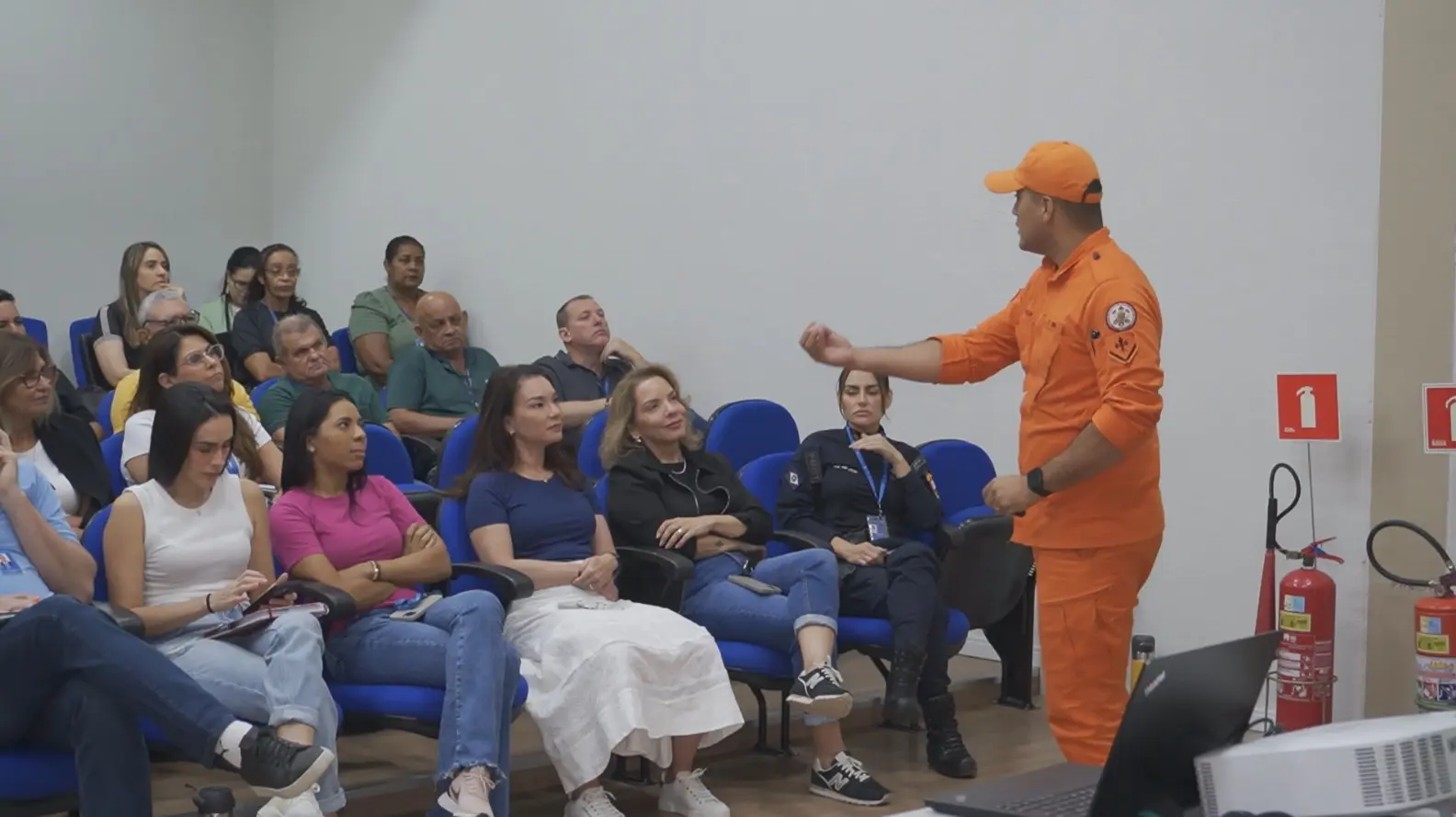 Foto horizontal em plano aberto que mostra cerca de 20 pessoas sentadas, assistindo a uma palestra de um bombeiro militar, que está em pé, com farda laranjada. 
