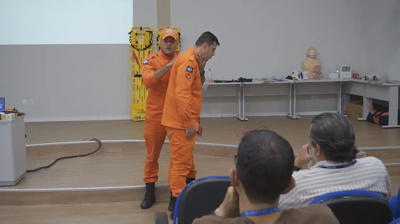 Foto horizontal em plano aberto que mostra dois bombeiros militares no centro de um auditório, demonstrando uma manobra de desengasgo. Na lateral direita da foto, aparecem dois homens sentados, assistindo à aula. 