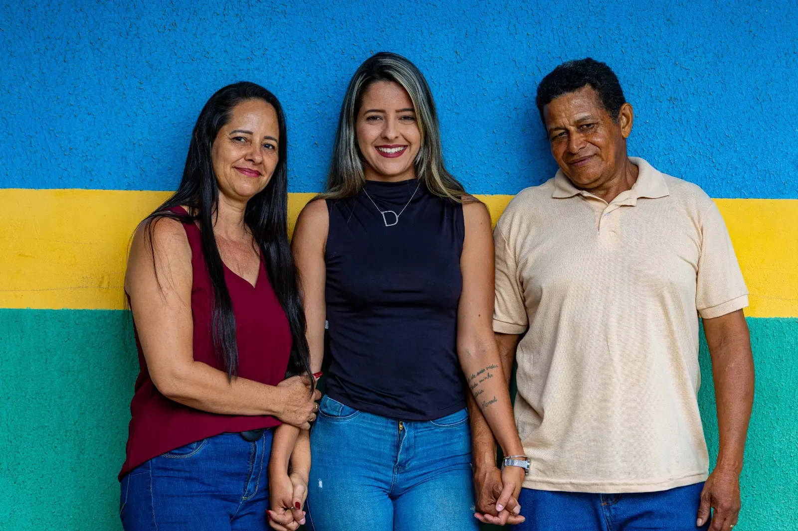 Em frente à parede colorida, três pessoas de mãos dadas sorriem para a câmera. A imagem transmite união, afeto e pertencimento, simbolizando o reconhecimento socioafetivo celebrado na expedição