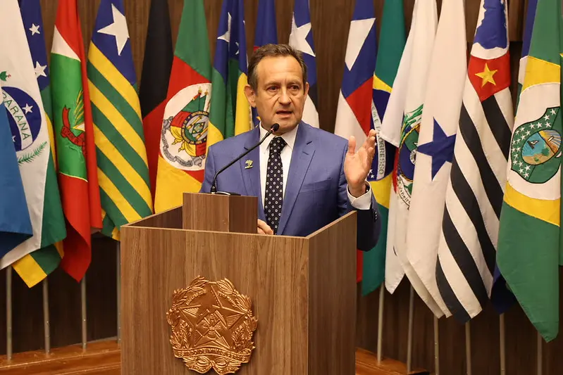 Presidente do TJMG, desembargador Corrêa Junior, fala na tribuna do Consepre. Ele é um senhor branco, de cabelo castanho, usando camisa branca, terno azul e gravata preta de bolinhas brancas.