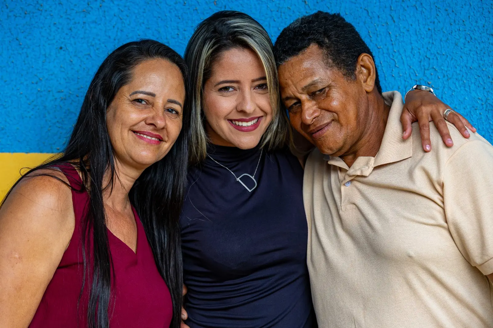 Três pessoas posam abraçadas diante de parede azul e amarela. A jovem ao centro sorri entre um homem e uma mulher que a abraçam com carinho, simbolizando afeto e reconhecimento familiar.