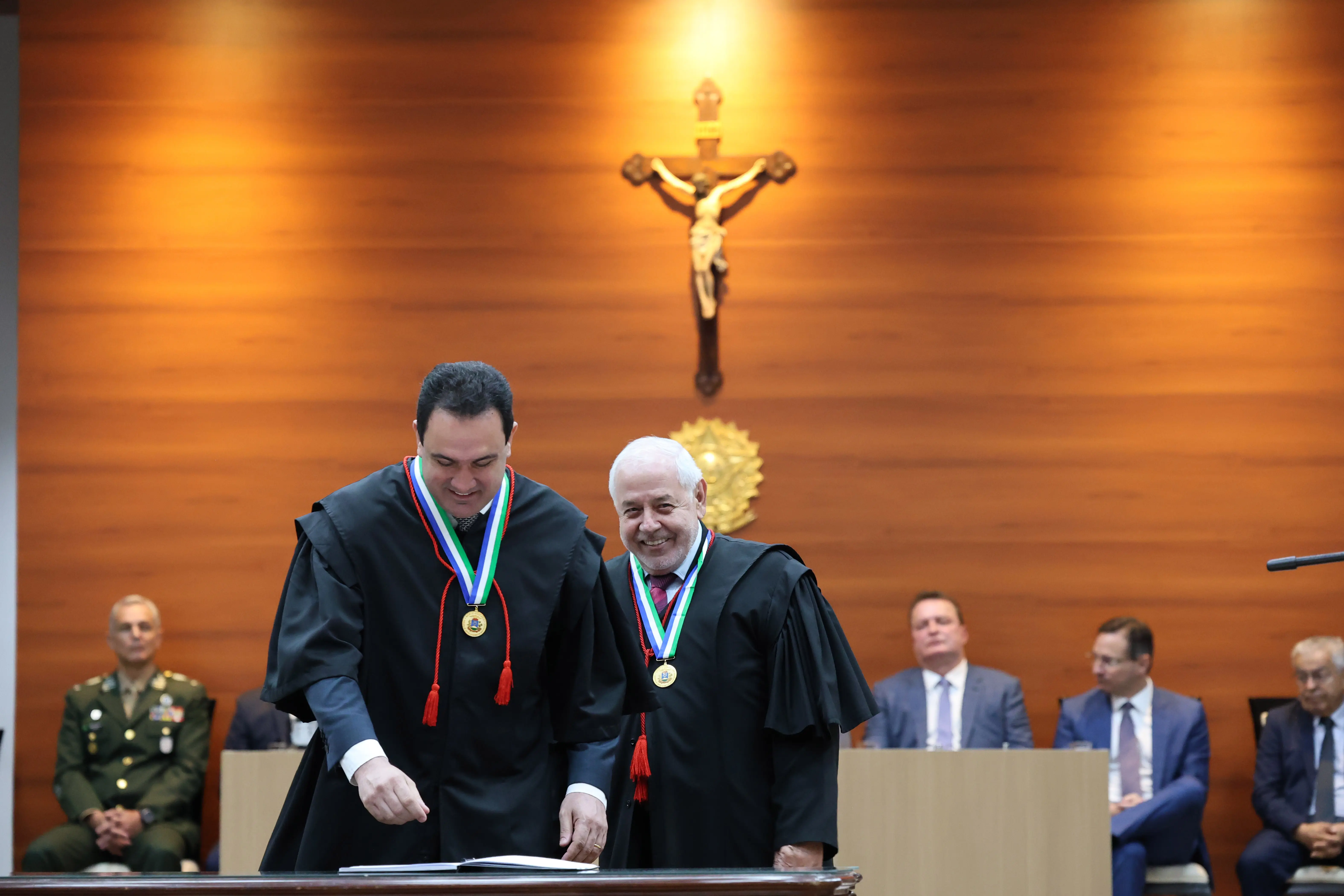 Desembargador Ricardo Almeida e presidente José Zuquim em pé, sorrindo, em frente à messa de assinatura do termo de posse. 