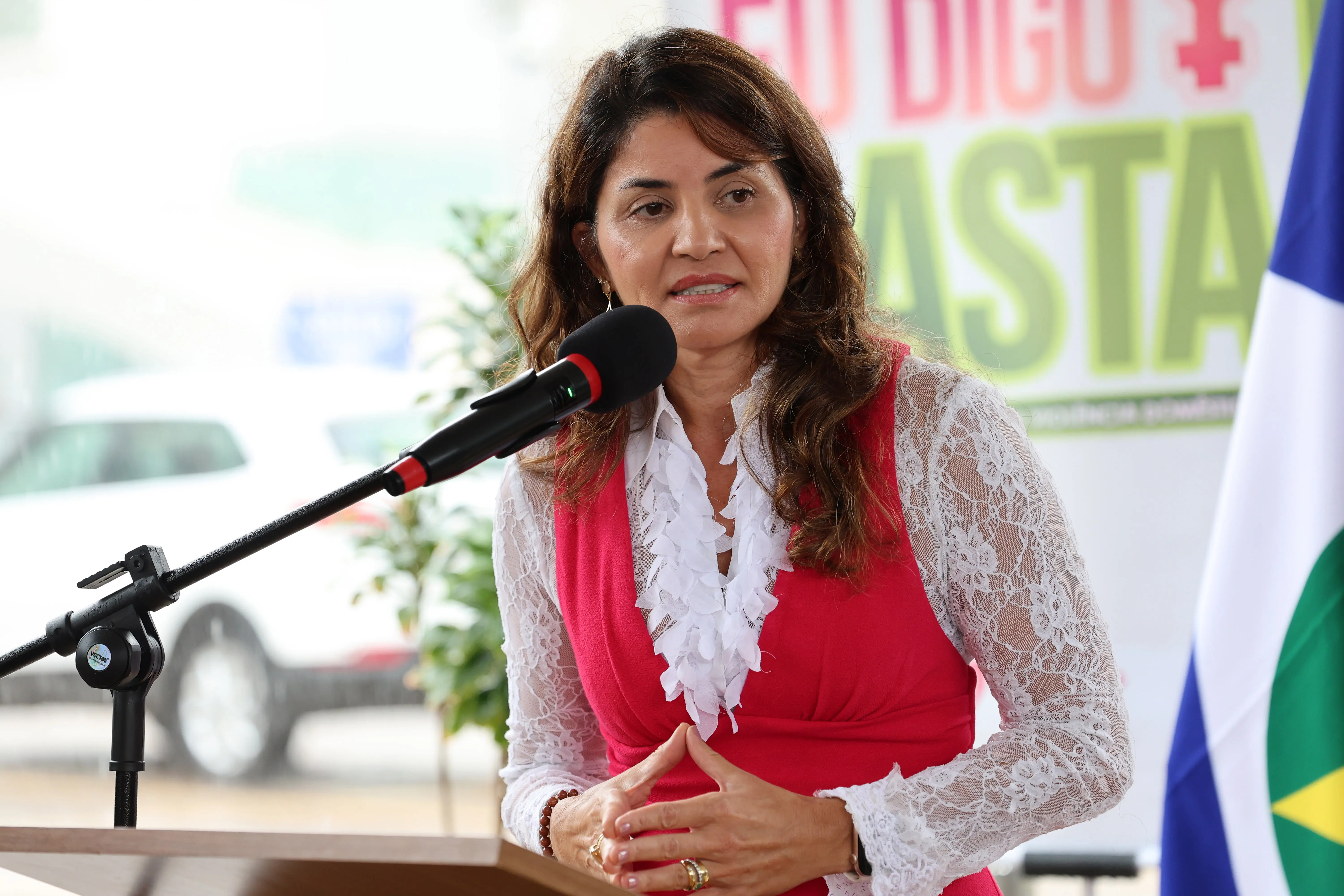 Maria Luziane Ribeiro de Castro, mulher de cabelos castanhos ondulados, fala ao microfone em um púlpito. Veste blusa branca rendada sob um colete rosa vibrante. Ao fundo, banner da campanha e bandeira.