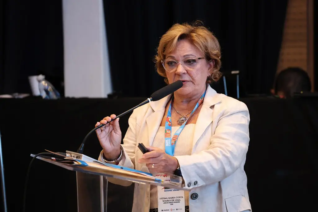 Foto horizontal que mostra a juíza do TJ do Rio Grande do Norte, Fátima Soares, falando ao microfone, no púlpito. Ela é uma mulher branca, de cabelos curtos e loiros, usando blusa branca, blazer branco, óculos de grau e crachá do Cojud.
