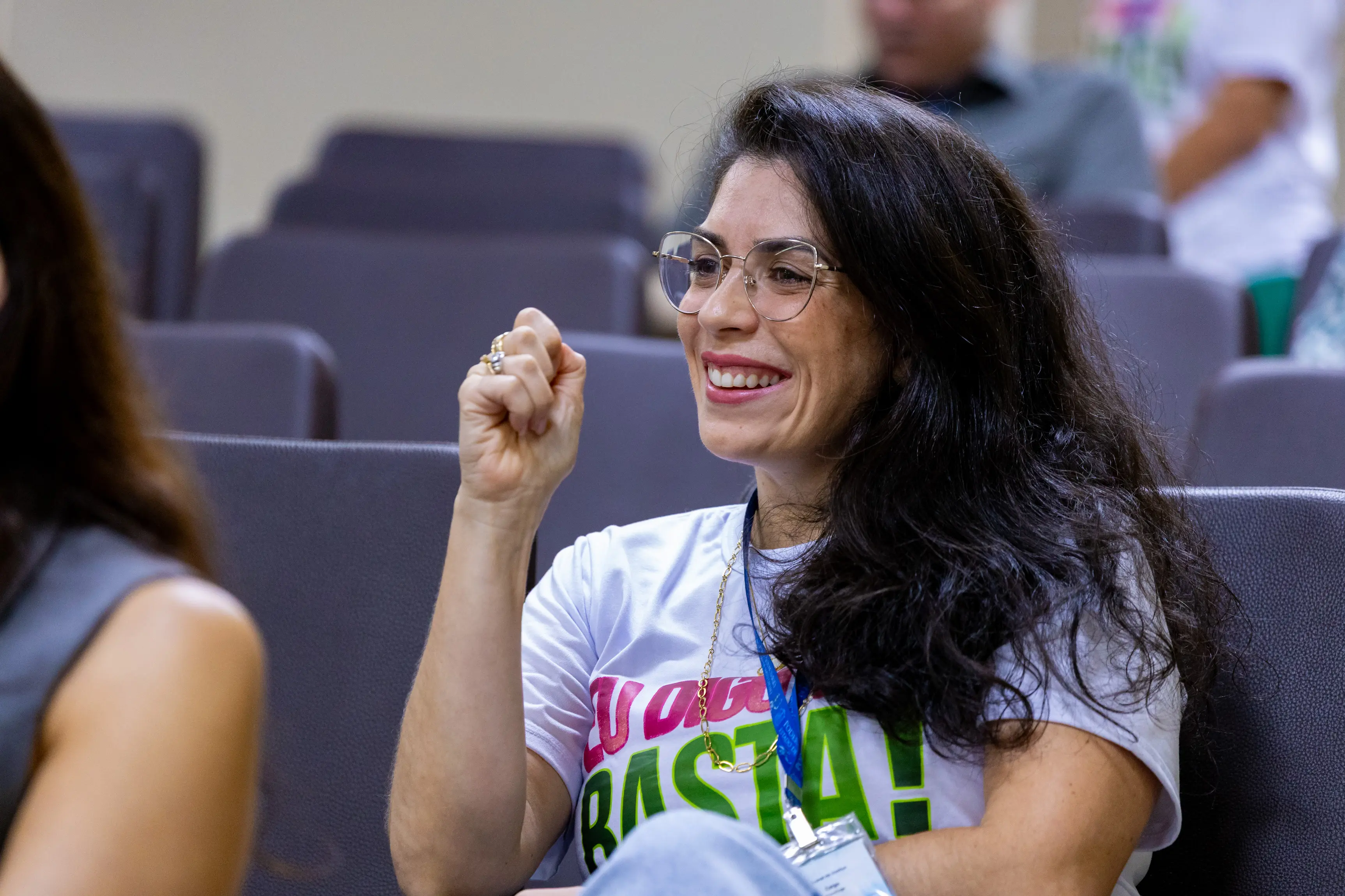 Mulher de cabelos escuros e camiseta branca com estampa colorida sorri enquanto levanta o punho em gesto de entusiasmo durante atividade no auditório. Ao fundo aparecem cadeiras do espaço do evento.