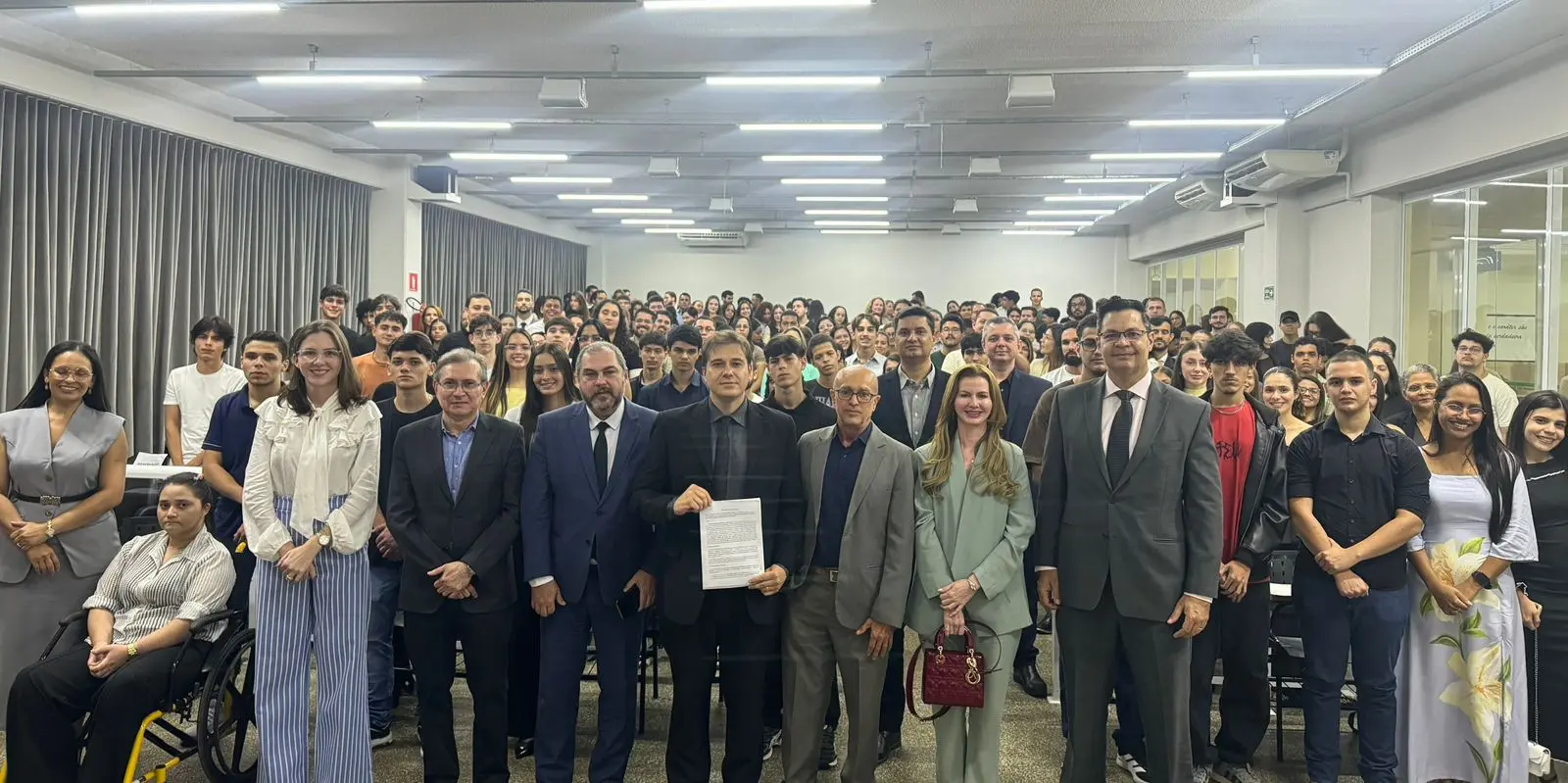 Autoridades e estudantes posam para foto coletiva em auditório após ato institucional. Ao centro, um magistrado segura documento assinado, enquanto dezenas de participantes ocupam o espaço ao fundo.