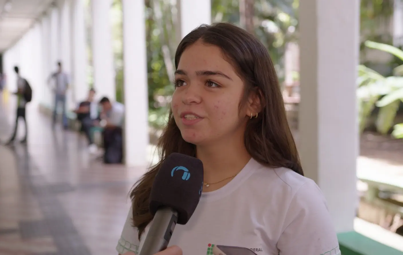 Foto horizontal em plano médio que mostra a estudante Daphny Pedraza durante entrevista à TV Justiça, no corredor do IFMT. Ela é uma adolescente de pele branca, cabelos longos, castanhos, lisos e soltos, olhos castanhos claros, usando camiseta branca da escola.