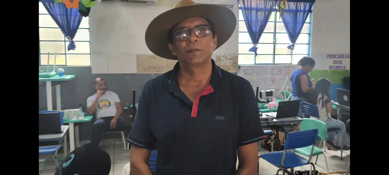 Homem de óculos e chapéu de abas largas veste camisa polo azul. Ele está em uma sala de aula com mesas, cadeiras azuis e pessoas ao fundo. O ambiente sugere um dia de atendimento ou evento social.